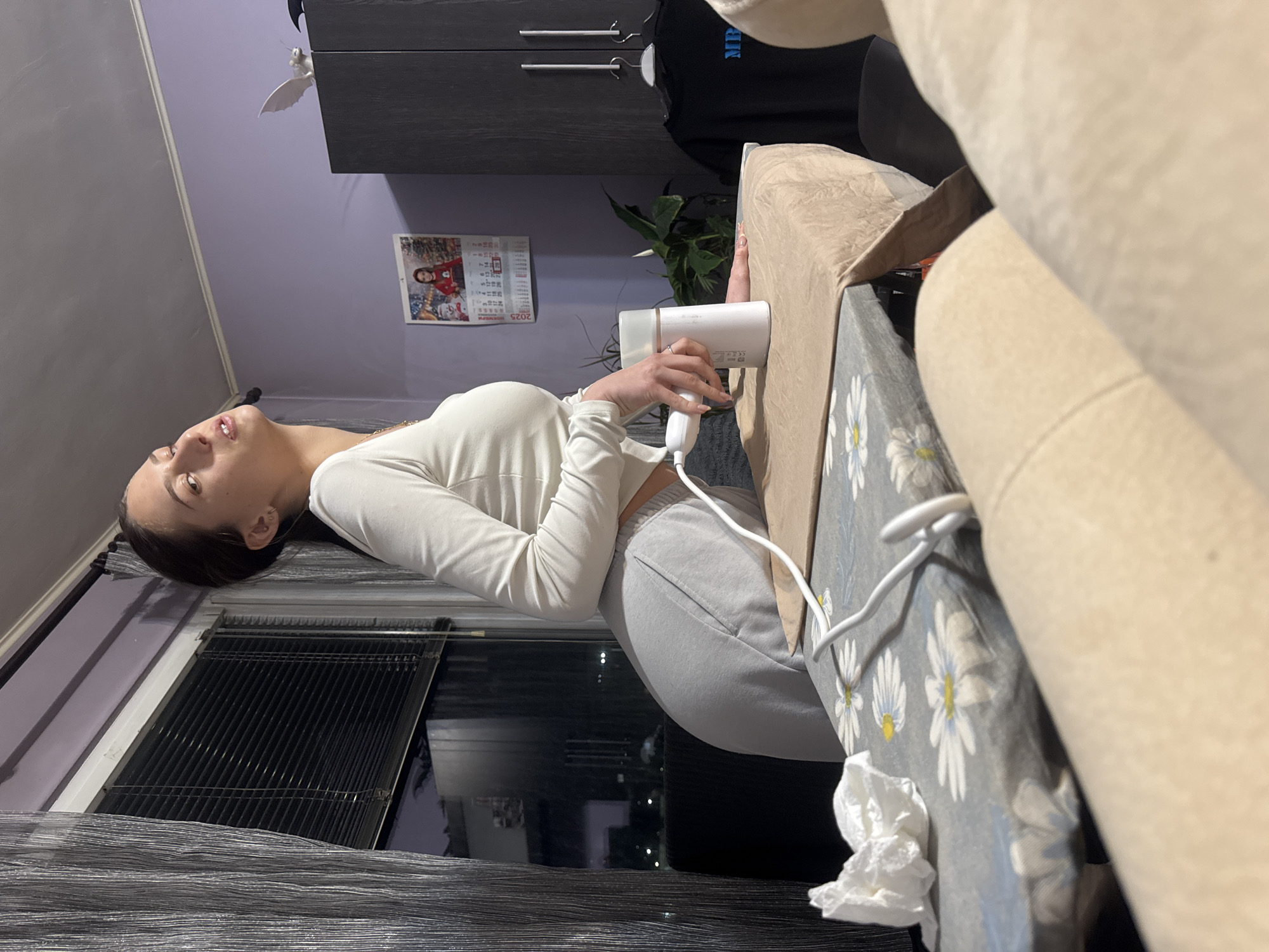 A woman is sitting on a couch, drying her hair with a white hair dryer. She is wearing a white long-sleeved shirt and gray sweatpants. The couch has a floral pattern and a beige cushion. The room is decorated with a calendar on the wall and a black cabinet. A window with closed blinds is visible in the background. The location is not identifiable as Sofia, Bulgaria.