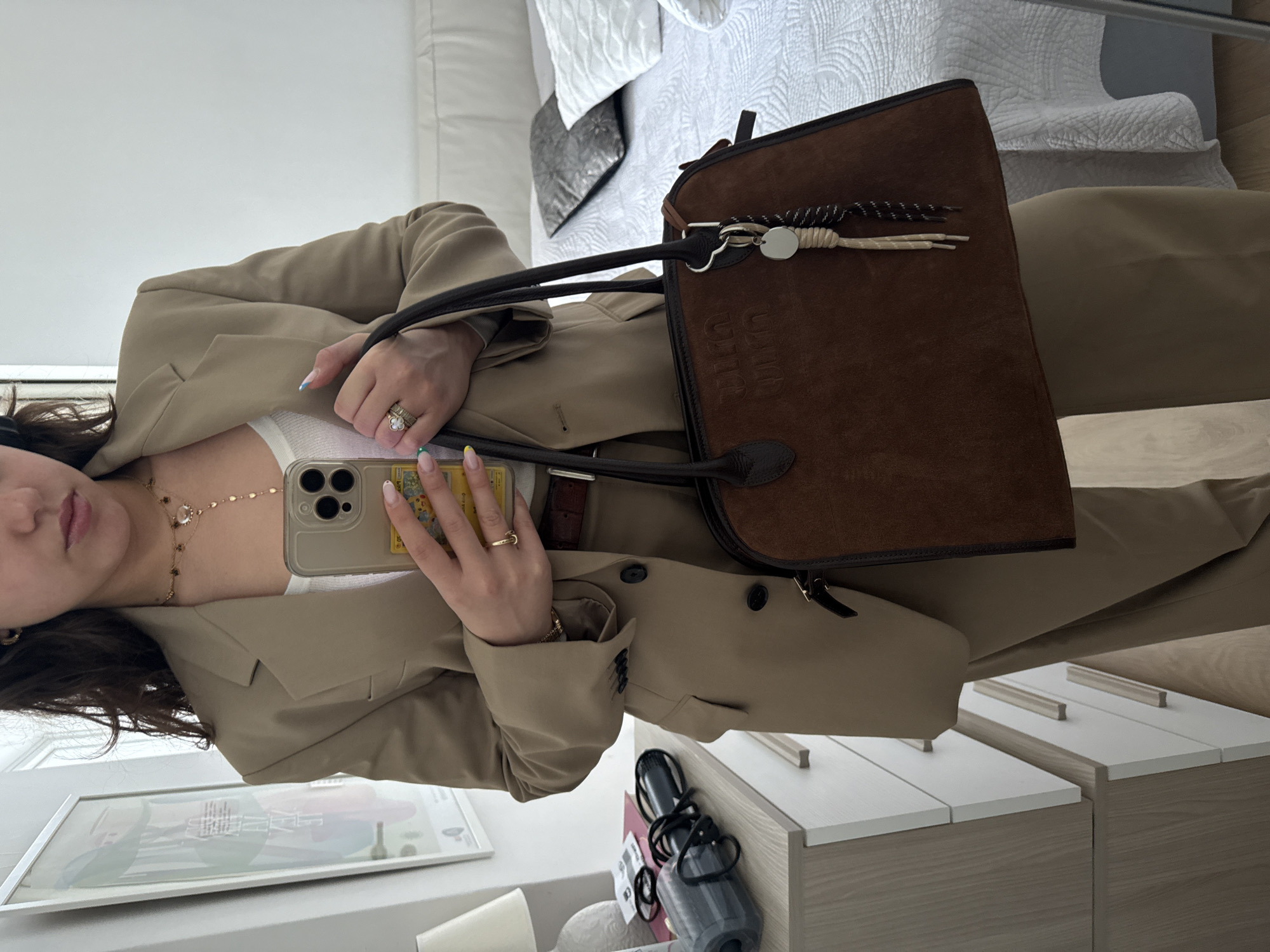 The image displays a mirror selfie of a person, likely a woman, captured indoors. The person is wearing a light beige or khaki blazer over a white ribbed camisole, with what appears to be matching trousers. She holds a light-colored smartphone with a transparent case that seemingly holds a card, possibly a Charizard Pokémon card with Japanese text. Her manicured nails feature light blue and yellow tips. On her right hand, she wears multiple rings, including a prominent one with a green four-leaf clover design and another with pearl-like accents. Several delicate gold necklaces adorn her neck.
She carries a dark brown suede tote bag with black leather handles and trim, conspicuously embossed with the brand name "LOEWE" across its surface. A decorative bag charm, comprising a silver disc and braided cords, is attached to one of the handles.
The background suggests a bedroom or dressing area. A neatly made bed with a white, patterned duvet cover and light grey pillows is visible. Below, two light-colored wooden nightstands or drawer units with white tops and light wooden handles are positioned. A framed picture or artwork hangs on a white wall, though its text is unreadable. Other items include a black hair dryer and a clear water bottle on the nightstand. The lighting indicates it is daytime.