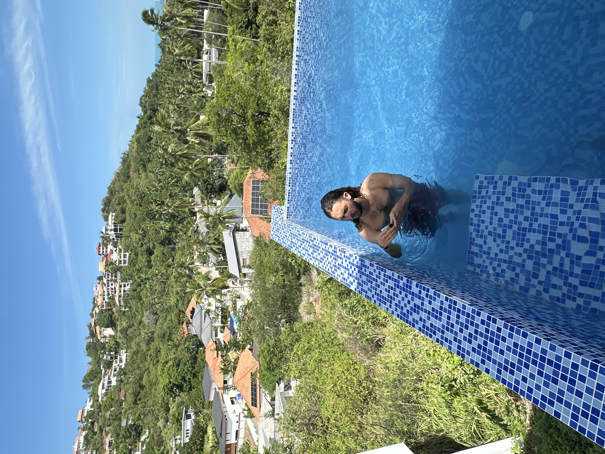 A man with long, dark hair stands in a blue tiled swimming pool, looking down at his hands. He is shirtless and wearing dark swimming trunks. The pool overlooks a hillside covered in lush green vegetation and houses, with palm trees scattered throughout. The sky above is a clear, bright blue with a few wispy clouds. The scene appears to be a residential area on the island of Ko Samui, Thailand, given the tropical foliage and architectural style of the houses.