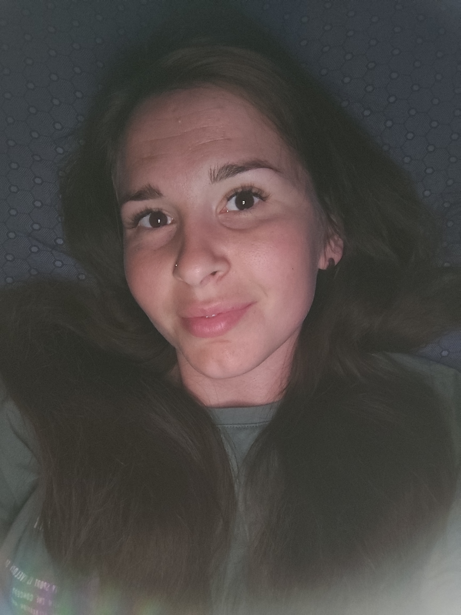 A close-up, overhead shot features a young woman lying down, looking directly at the camera. Her long, dark brown hair spreads out around her head against a dark blue, patterned surface. The woman has fair skin, dark eyebrows, and dark eyes. She is wearing a green t-shirt. The lighting appears to be artificial, casting a slight reddish hue on her face. There is no discernible background information that would indicate a specific location within the city of Fokino, Russia.