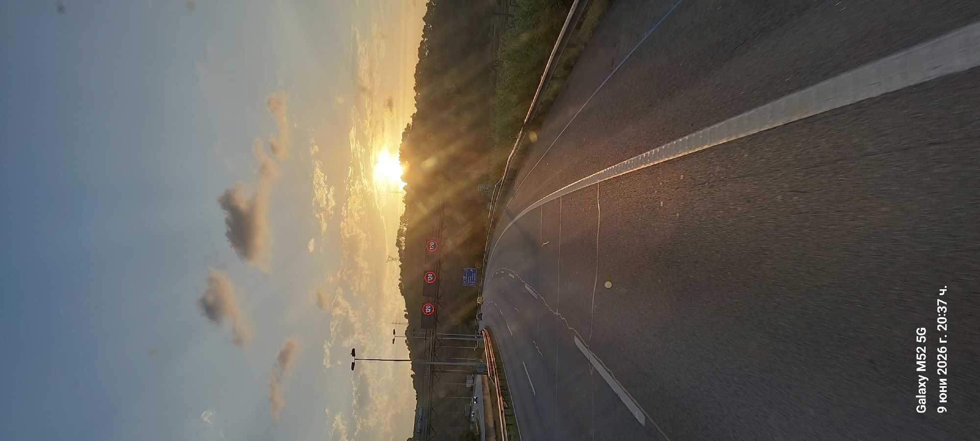 The image is a photograph of a highway at sunset, viewed from the perspective of a moving vehicle. The sun is setting behind a tree-covered hill, casting long shadows and lens flares across the scene. Multiple circular speed limit signs showing "120" are visible on an overhead gantry. The road has multiple lanes and is marked with white lines. In the lower left corner of the image, text reads "Galaxy M52 5G 9 юни 2026 г. 20:37 ч.", indicating the device used and the date and time the photo was taken, which is in Bulgarian. The location is identified as Castellbisbal, Spain. There are no people visible in the image.