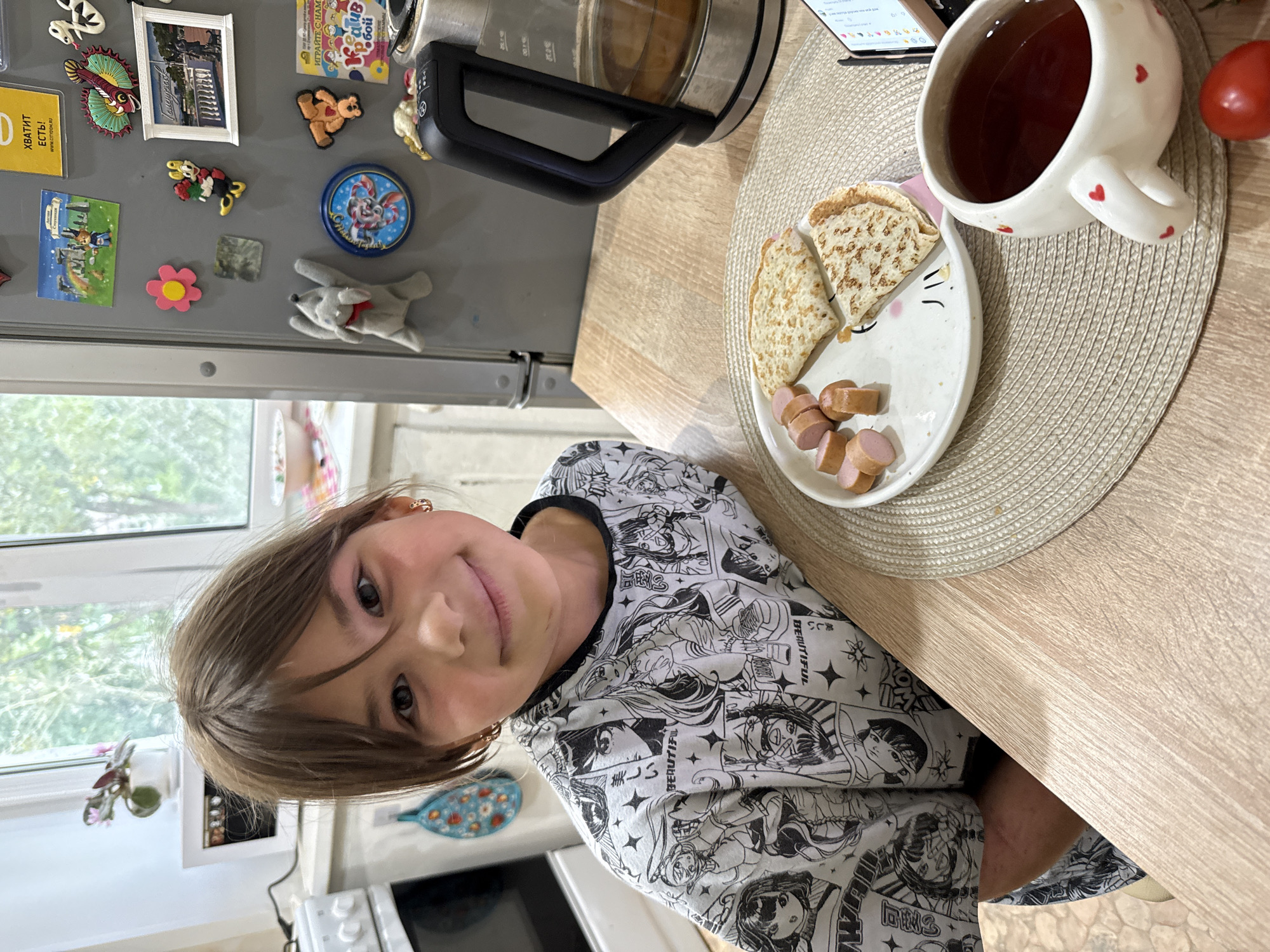 In a domestic kitchen setting, a young girl with dark hair and brown eyes sits at a wooden table. She is wearing a black and white t-shirt with a manga-style print. In front of her is a plate with sliced sausages and two folded crepes. A white mug decorated with small red hearts, filled with dark liquid, sits beside the plate. A kettle is visible in the background on the right, and a phone rests on the table to the right of the mug. The kitchen wall features a refrigerator adorned with various magnets and pictures. The scene is illuminated by natural light filtering through a window. The image is not situated in the city of Rusanovo, Russia.