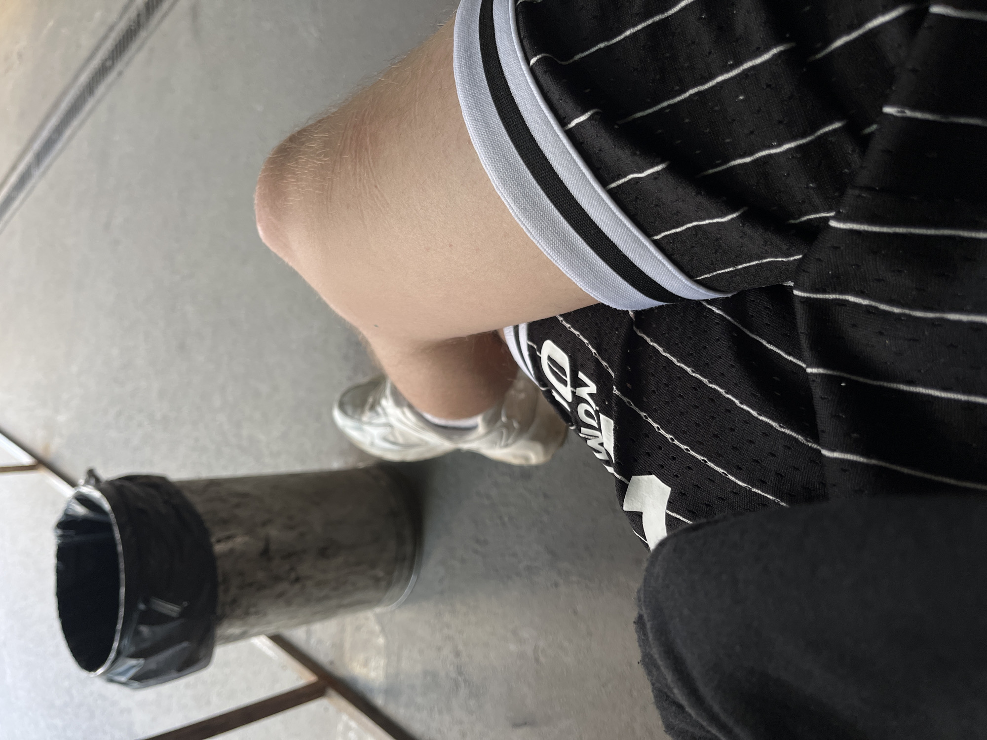 The image shows a close-up, overhead view of a person's legs and feet, wearing black athletic shorts with white pinstripes and white athletic shoes. The person is sitting down on a grey, textured surface, possibly concrete. To the left of the frame, there is a metal cylinder covered with black tape around its opening, which appears to be a trash can or some kind of industrial pipe. The lighting suggests it is daytime, and the setting appears to be outdoors or in an open-air industrial area. The overall mood is neutral, with no strong emotional cues. There is no visible text in the image.