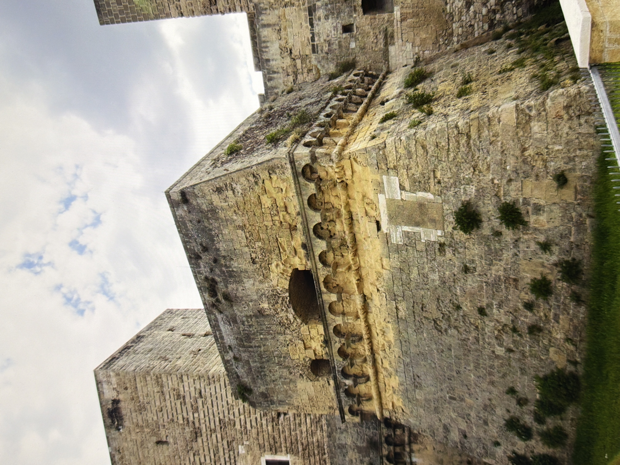 The image shows a close-up, low-angle view of a large, ancient stone fortress. The fortress is constructed from light-colored, weathered stone blocks of varying sizes. The main structure in the foreground is a multi-layered fortification with a prominent overhanging section adorned with a series of decorative arches. Above this arched section, there are two small, dark openings that appear to be windows or arrow slits. The stone walls are partially covered in patches of moss and small green plants, indicating age and exposure to the elements.
To the left of the main structure, a taller, more angular stone tower is visible, also constructed from large, rough-hewn blocks. Its surface is textured and shows signs of significant wear. The sky above is partly cloudy, with patches of white and gray clouds against a lighter sky.
The bottom of the image shows a strip of bright green grass and a metal fence. The overall impression is of a historic and formidable military structure.
The image does not show the city of Kyiv, Ukraine.