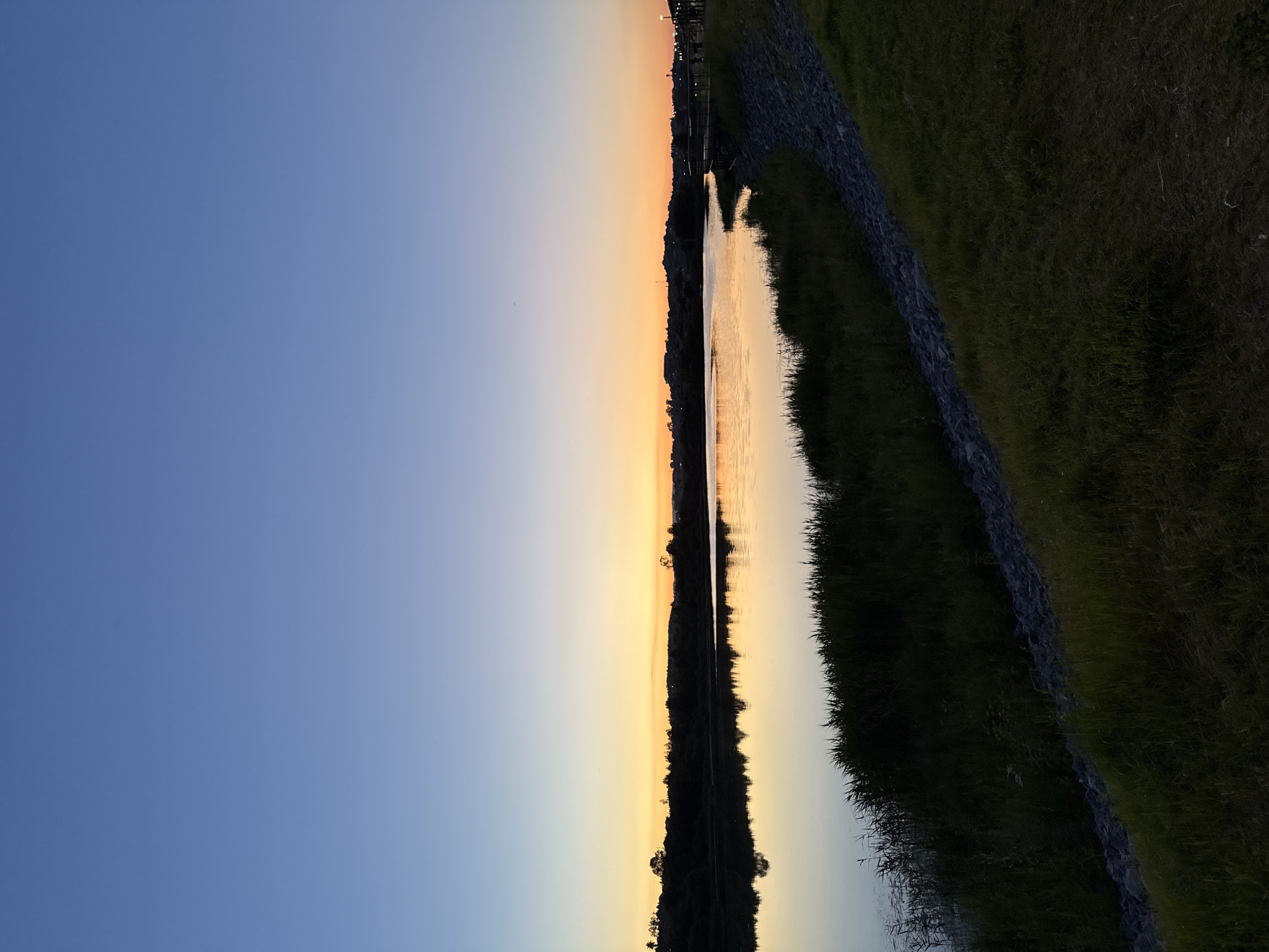 The image depicts a serene lake at dusk, with the sky transitioning from a deep blue to a soft orange hue near the horizon. Silhouetted trees and a distant treeline form the backdrop across the lake. The water's surface is calm and reflective, mirroring the sky and the dark vegetation along its edge. Tall grasses and reeds grow along the immediate bank, giving way to a grassy slope dotted with rocks. In the distance, on the right side of the frame, a boardwalk or pier with lampposts is visible, suggesting a developed area. The overall scene evokes a peaceful, natural setting during twilight. The location is described as being in the city of Miass, Russia.