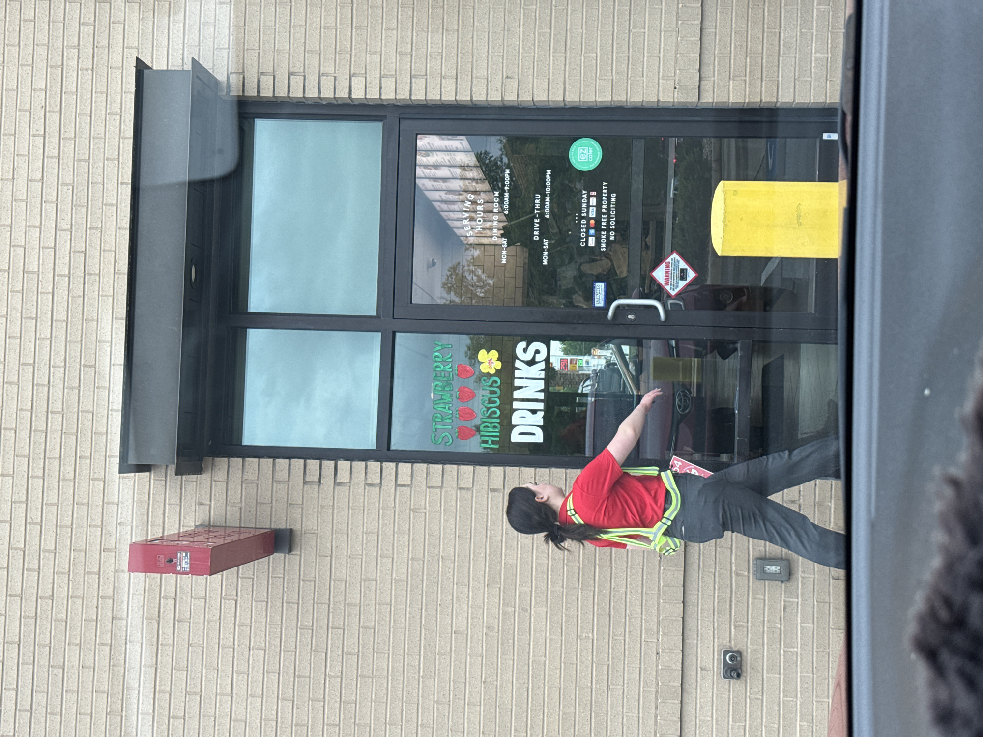 Here is a description of the image:
The image shows the exterior of a building, likely a restaurant in Mountain Park, United States of America. The building has a brick facade with a dark-framed glass door. On the door are several signs, including one advertising "Strawberry Hibiscus Drinks," another displaying the restaurant's operating hours (6:00 AM to 9:00 PM for dining room and 6:00 AM to 10:00 PM for drive-thru, Monday to Saturday) and indicating it's closed on Sundays. Other signs state "Smoke Free Property" and "No Soliciting."
A person wearing a red shirt and a safety vest is visible in front of the building. The person is facing away from the camera. A red box is fixed on the wall of the building to the left of the door. An electrical outlet or similar fixture can be seen on the wall to the right of the door, above a possible security camera.
The overall impression is that it's a daytime scene at a fast-food or casual dining establishment.