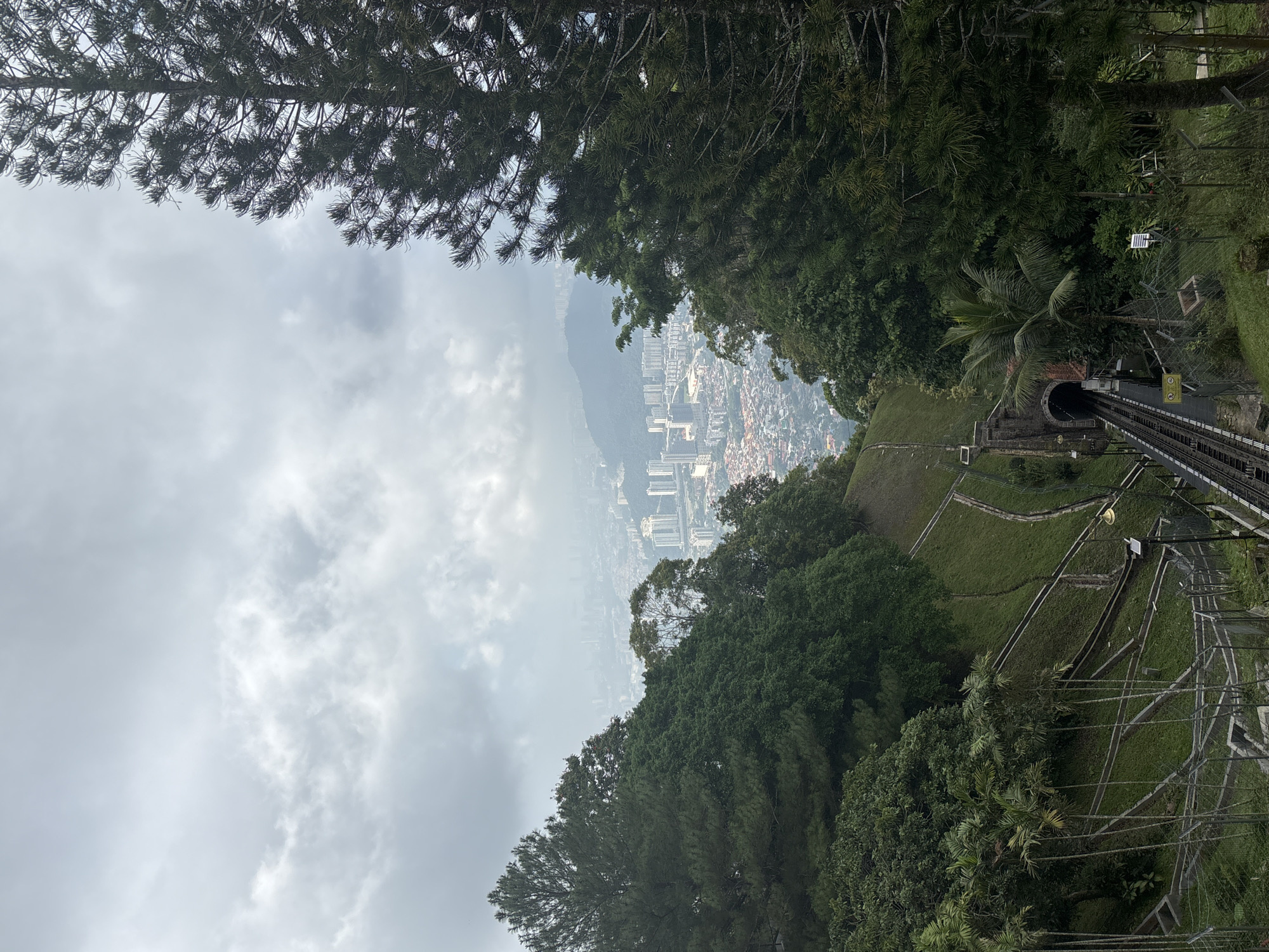 This is a photograph taken from a high vantage point, looking down a steep slope towards a city and the ocean beyond. In the foreground, a funicular railway track descends into a tunnel carved into the hillside. Dense green foliage, including palm trees and tall evergreen trees, borders the railway on both sides.
The slope itself is covered in neatly manicured grass, with visible terracing and retaining walls that suggest a park or garden area. The railway tracks are dark metal, with the characteristic gauge of a funicular system. A single lamp post with a yellow light is visible to the right of the tracks.
Beyond the immediate greenery, the city of George Town, Malaysia, sprawls out below. A mix of modern high-rise buildings and older, lower-rise structures are visible, interspersed with roads and what appear to be more patches of green space. The city is hazy, suggesting it is either distant or that there is atmospheric moisture. In the far distance, a hazy line indicates the presence of the ocean.
The sky is overcast with thick, gray clouds, which diffuse the light and create a somewhat muted color palette for the entire scene. There are no people or animals visible in the photograph. The image captures a transition from a lush, natural environment to an urban landscape, framed by the dramatic slope and the descending railway.