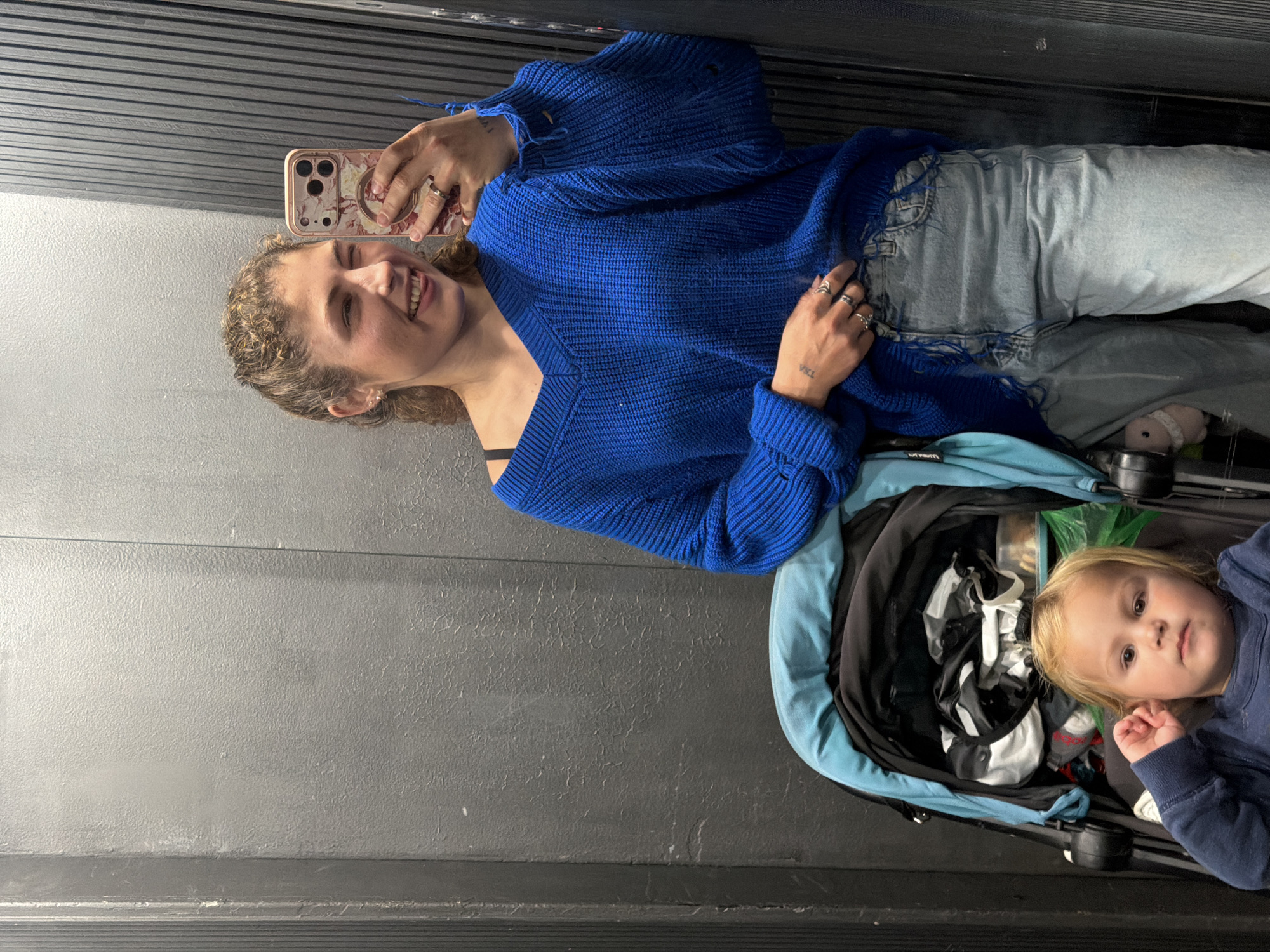 A fair-skinned woman with curly hair is taking a selfie with her phone in an elevator. She is wearing a blue sweater and jeans, and has several rings on her fingers. A small child with blonde hair lies in a stroller next to her. The elevator walls are gray. The location is unknown and cannot be determined.