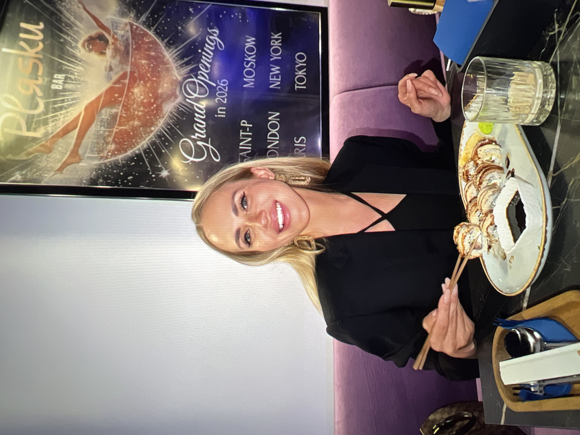 A woman with blonde hair and a black blazer is seated at a table, holding chopsticks and eating sushi. A plate of sushi, a glass of water, and cutlery are on the table. Behind her is a purple couch and a poster with text including "Plasku BAR" and "Grand Openings in 2026". The setting appears to be a restaurant. There is no information available to suggest the location is in the city of Unknown, Russia.