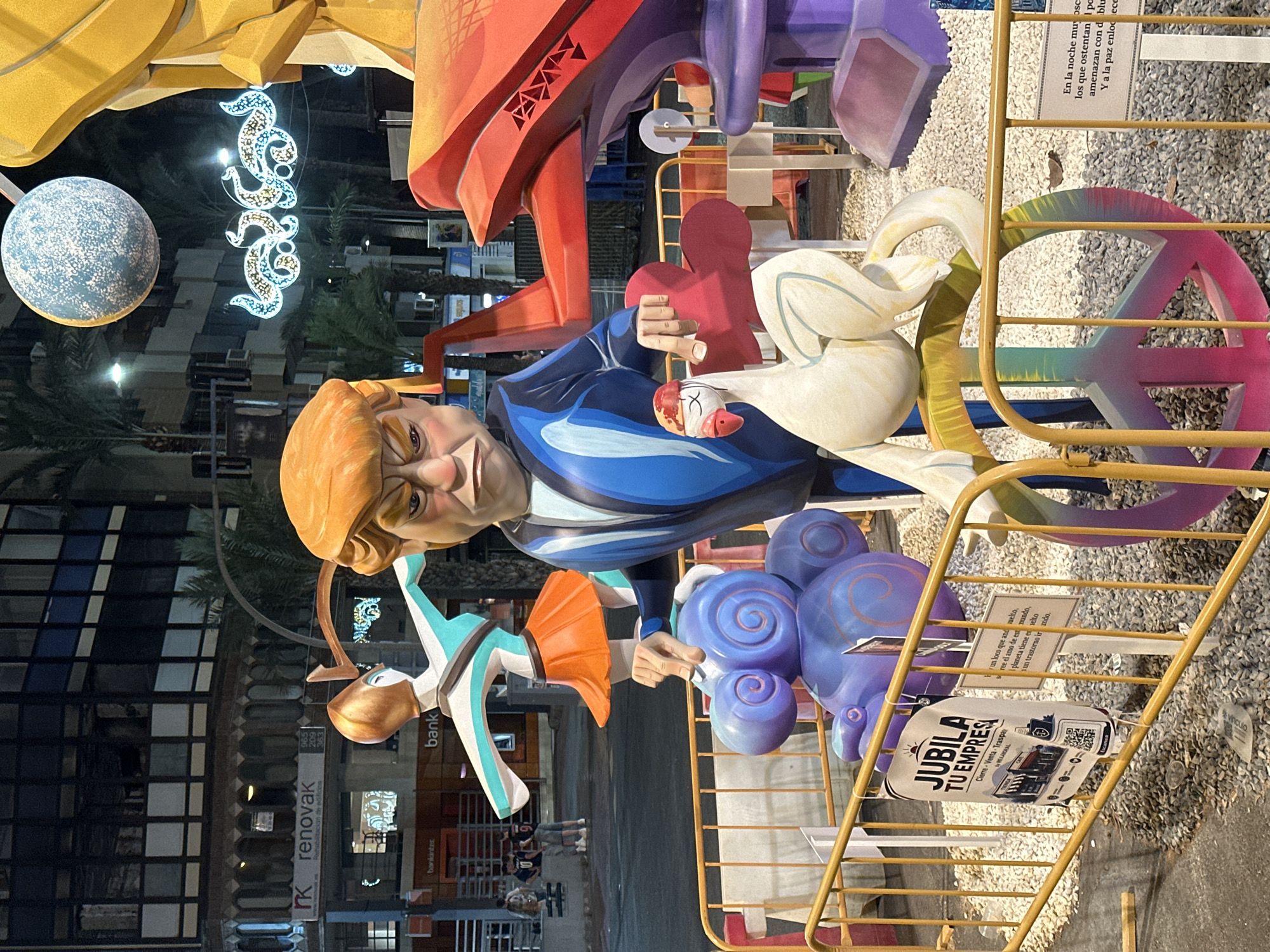 The image depicts a street scene at night in Alicante, Spain, featuring a large-scale art installation, likely part of a festival or parade. The central figure is a caricature resembling Donald Trump, depicted with an angry expression, wearing a blue suit. He is holding a red object in his right hand and pointing with his left index finger towards a purple, cloud-like sculpture. To the left of Trump, a stylized figure in a teal and orange costume is in motion, with arms outstretched. To the right of the main figure, a white chicken-like sculpture with a red headdress is perched atop a large, colorful peace sign that extends from the foreground. The art installation is cordoned off by yellow barriers, with a gravel-covered ground. Signs are attached to the barriers, including one with the text "JUBILA TU EMPRESA." In the background, buildings with illuminated windows and palm trees are visible, suggesting an urban setting. Decorative lights, including a glowing orb and a serpentine design, are strung across the scene, adding to the festive atmosphere. Several people are present in the background, observing the display.