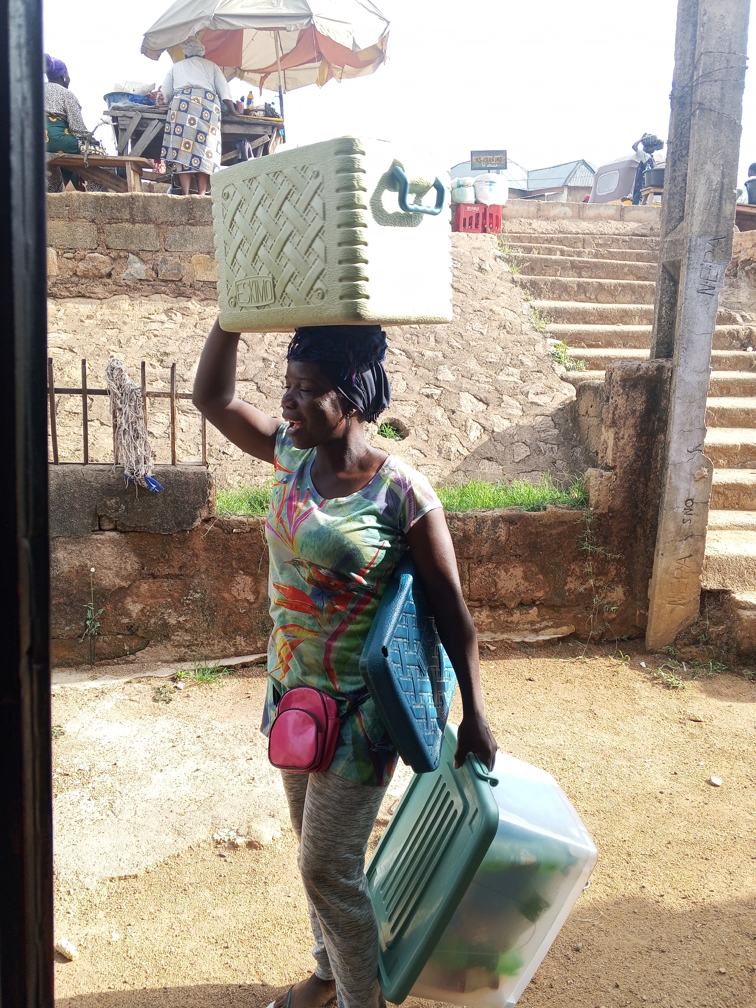 A woman in Unknown, Unknown carries two plastic containers and a cooler on her head. She wears a floral top, gray pants, and sandals. A pink bag is slung over her shoulder. She stands on a dirt path, with a stone wall and steps leading upwards in the background. A vendor operates a food stand atop the wall. The container on her head is marked "ESKIMO."