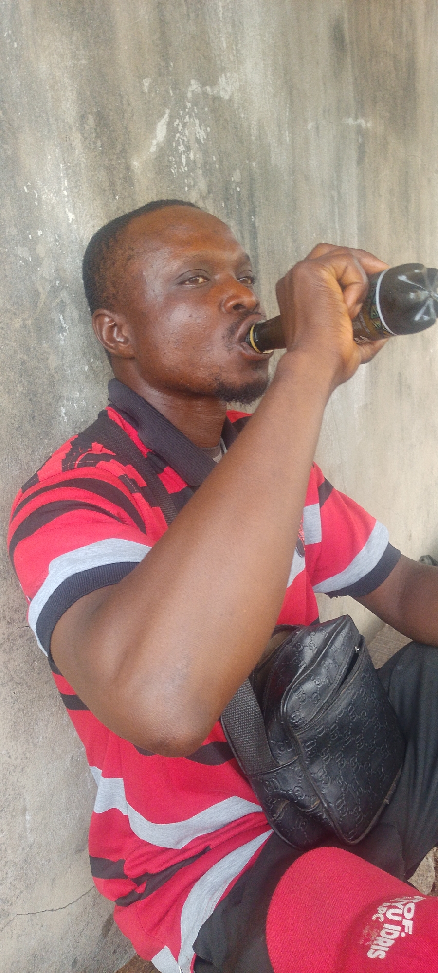 A man is drinking from a brown bottle, sitting against a concrete wall. He is wearing a red and black striped shirt with gray accents, black pants, and red undergarments with white lettering, with a black shoulder bag on his lap. The wall appears to be exterior and is rough, possibly aged concrete. The location is Lokoja, Nigeria, but further details about the specific area are not visible.