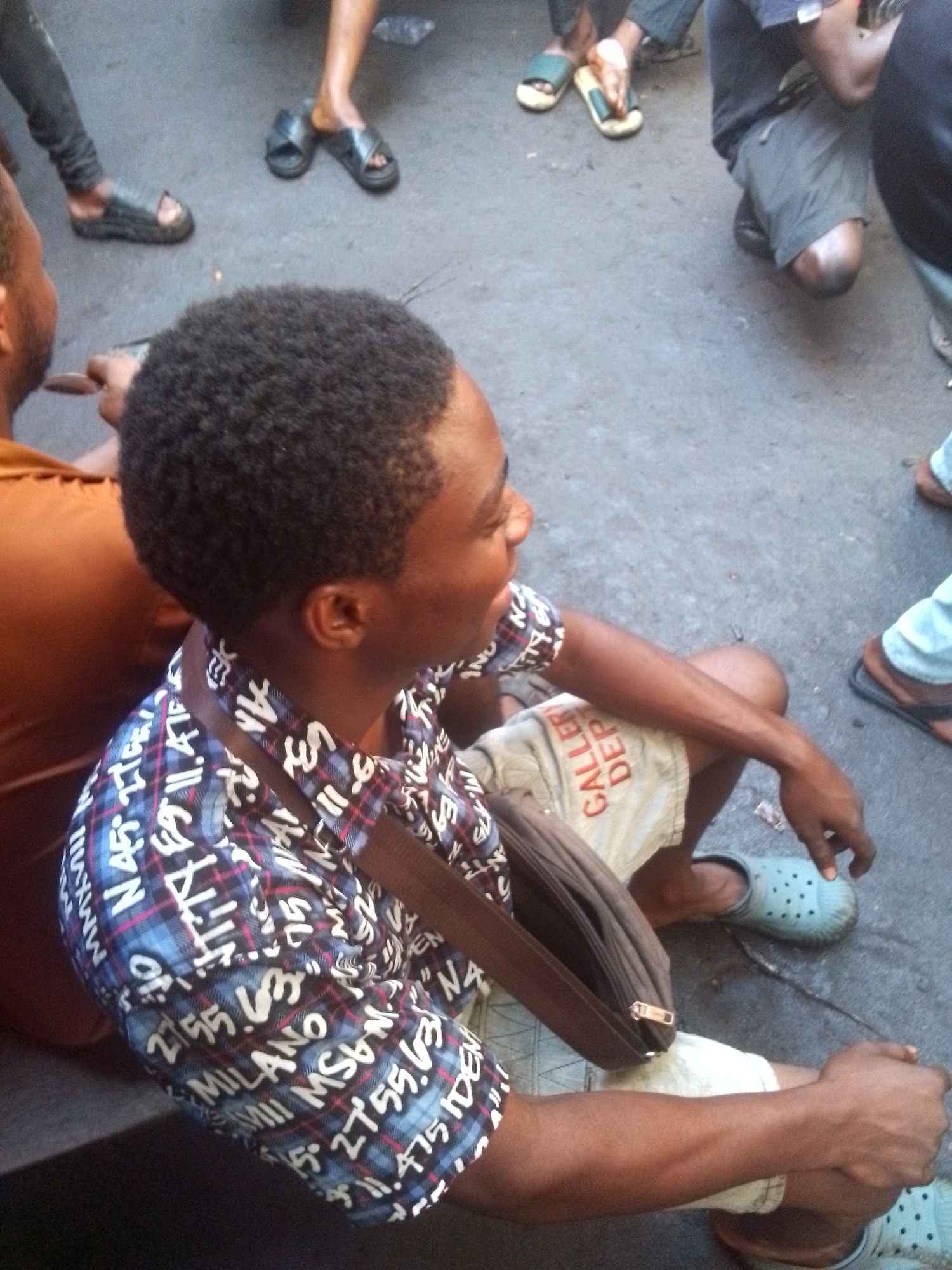 The image shows a man sitting on the ground amidst a gathering of people. The location appears to be a city street, likely in Onitsha, Nigeria, given the context. The man is wearing a patterned shirt with the word "Milano" and other text, khaki shorts with "GALLER DEP" written on them, and blue clog-style sandals. He has a brown shoulder bag. Other people are visible, mostly from the knees down, wearing various types of footwear. The ground is concrete.