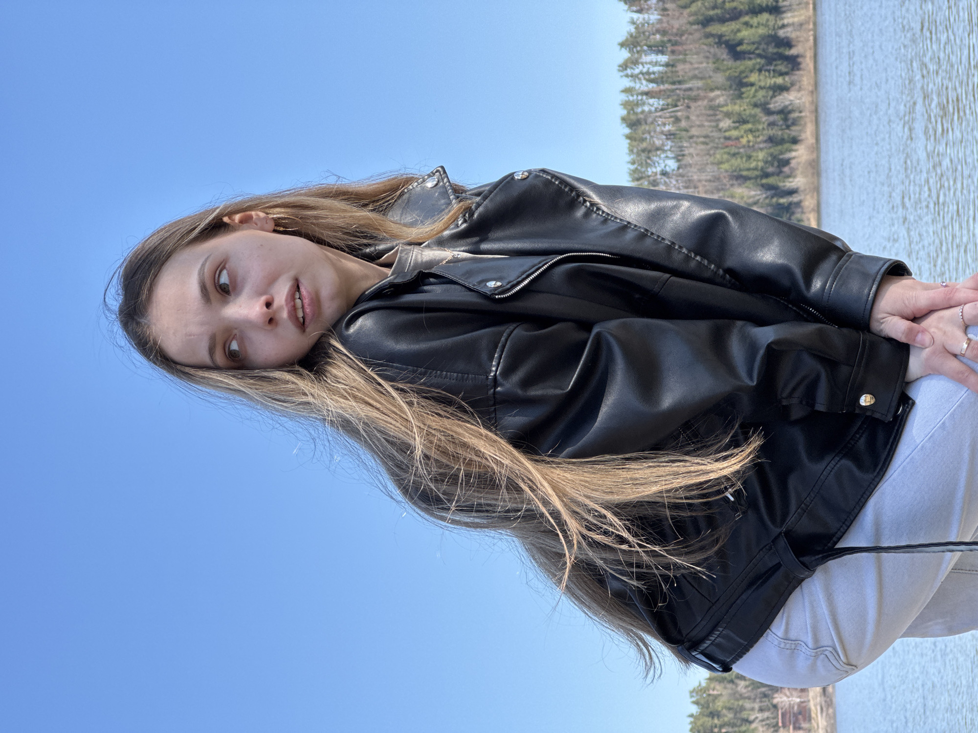 A young woman with long, light brown hair is seen in an outdoor setting. She is wearing a black leather jacket and white pants. Her hands are clasped together. The background consists of a body of water and trees. The sky is clear and blue. The image is oriented on its side.