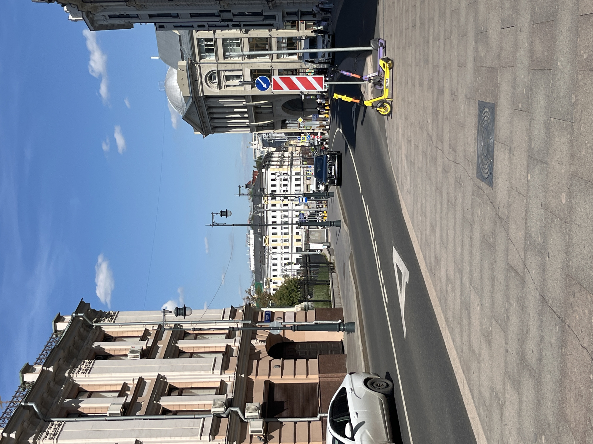 The image depicts a street scene in Moscow, Russia, likely on a sunny day with a clear blue sky dotted with a few white clouds. The architecture suggests a historical district with ornate, light-colored buildings lining both sides of the street.
On the left, a multi-story building features classical architectural elements, including columns, decorative moldings, and arched windows. Air conditioning units are visible on its facade. A white car is parked along the curb.
The street itself is paved and has lane markings, including a white arrow pointing forward. A black sedan is driving down the street.
On the right side of the street, a large, light-colored building with a domed roof and classical columns stands prominently. Nearby, a white van is parked. Several people are visible walking on the sidewalk in the distance.
In the foreground on the right sidewalk, two electric scooters are parked. One is yellow, and the other is purple. A traffic sign with red and white stripes and a blue circular sign with a white arrow is also present.
The overall impression is of a bustling urban environment with a blend of historical and modern elements.