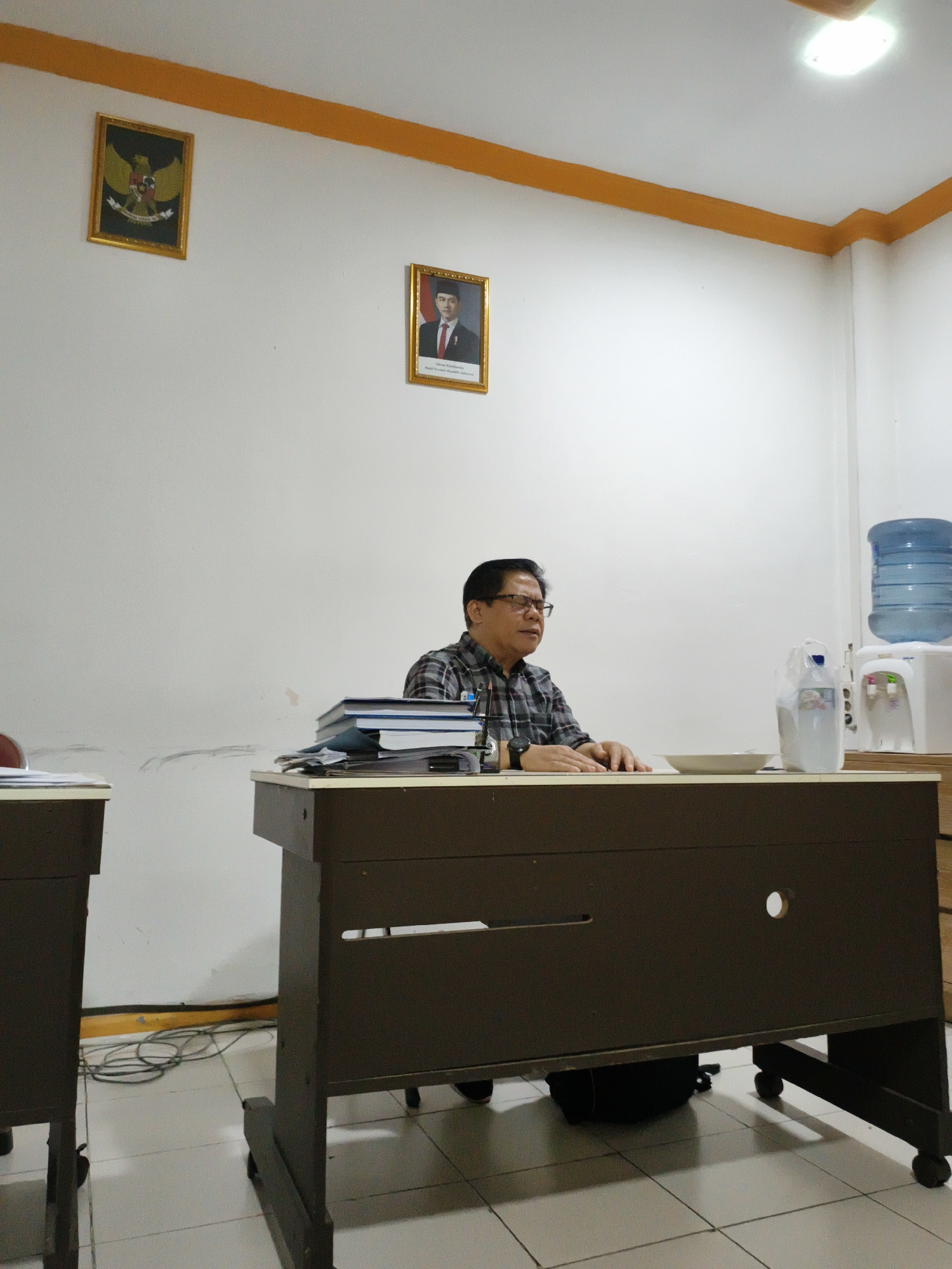 A middle-aged man with glasses and a plaid shirt is seated at a dark brown office desk, facing slightly to his right, in what appears to be a meeting or work room in Cianjur, Indonesia. His hands rest on the desk, and he seems engaged, possibly listening or contemplating.
The room features white walls with a distinctive yellow trim running along the top perimeter and a white tiled floor. A fluorescent light fixture illuminates the space from the ceiling. On the wall, two framed pictures are prominently displayed. To the left is a golden-framed image of the Garuda Pancasila, the national emblem of Indonesia. To the right, another golden-framed portrait depicts H. Muhammad Jusuf Kalla, identified by the text below his image as "Wakil Presiden Republik Indonesia" (Vice President of the Republic of Indonesia).
On the man's left side of the desk, a stack of books and papers is neatly piled. To his right, a light-colored ceramic bowl, a large clear plastic water bottle, and a white water dispenser with a large blue jug are visible. Another desk is partially seen to the left, and electrical cords are coiled on the floor between the desks. A dark backpack is tucked away under the man's desk. The setting suggests an official or semi-formal institutional environment, likely during daytime given the bright indoor lighting.