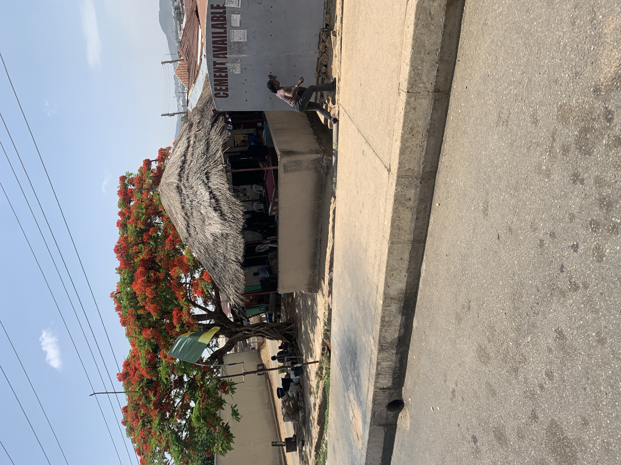 The image shows a street scene in Jos, Nigeria. A building with a thatched roof overhangs the sidewalk. A sign on the building reads "CEMENT AVAILABLE." A tree with red flowers is in front of the building. A person is walking along the sidewalk in front of the building. Other people are seated inside the building. The street is paved. Power lines are visible overhead. A green and yellow flag hangs from the tree. There are a few black plastic bags next to the tree.