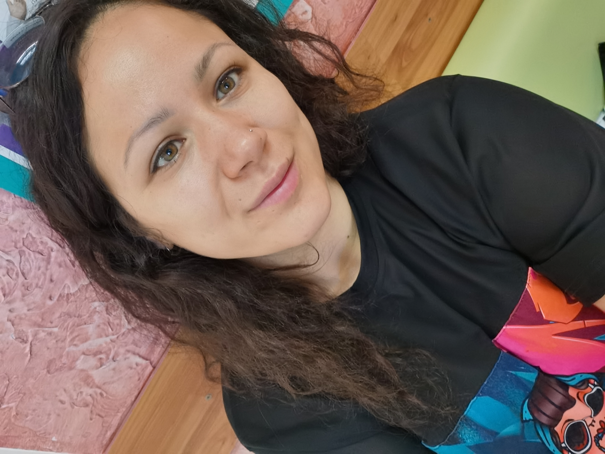 A close-up, angled selfie of a woman. She has light skin, dark wavy hair, and hazel eyes. A small stud is visible in her left nostril. She wears a dark-colored shirt with a portion of a colorful graphic visible on the lower right side. The background includes a pink wall with a textured surface on the lower left, wood paneling, and a portion of a light green wall on the upper right. The location is in the city of Chaikovsky, Russia.