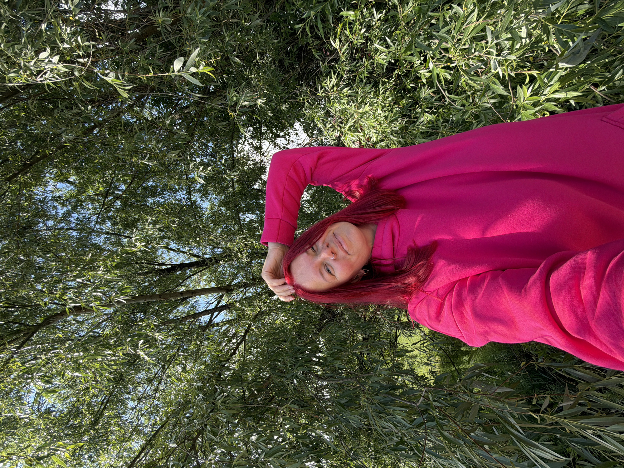 A person with vibrant red hair, wearing a bright fuchsia pink long-sleeved top or hoodie, is shown from a high-angle perspective, appearing to lie down outdoors. The image is rotated 90 degrees clockwise, making the person appear horizontal. They are looking towards the camera with a relaxed, slightly smiling expression, and one hand is resting near their temple.
The background is filled with a dense canopy of green leaves, likely from trees such as willows, with patches of clear blue sky visible through the foliage. The lighting suggests it is a bright, sunny day. The scene depicts a serene outdoor setting, possibly a park or natural area in Chaikovsky, Russia. There is no visible text in the image.