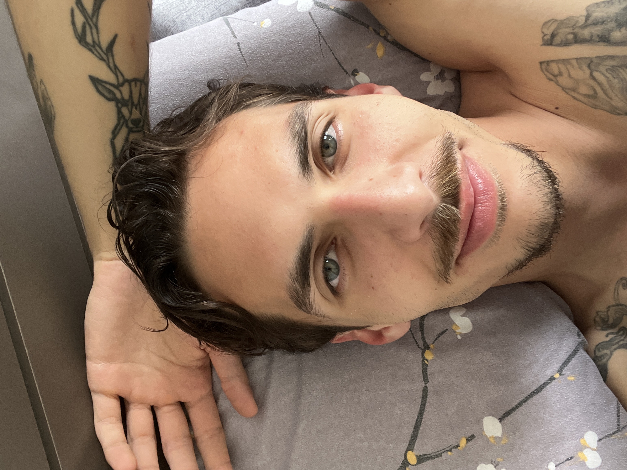 A man lies on a bed in an interior setting. He is fair-skinned with brown hair, blue eyes, and a mustache. The man has tattoos on both arms, one featuring a deer. He is lying on a gray pillow with a floral pattern. No specific details indicate that the location is Rafailovići, Montenegro.