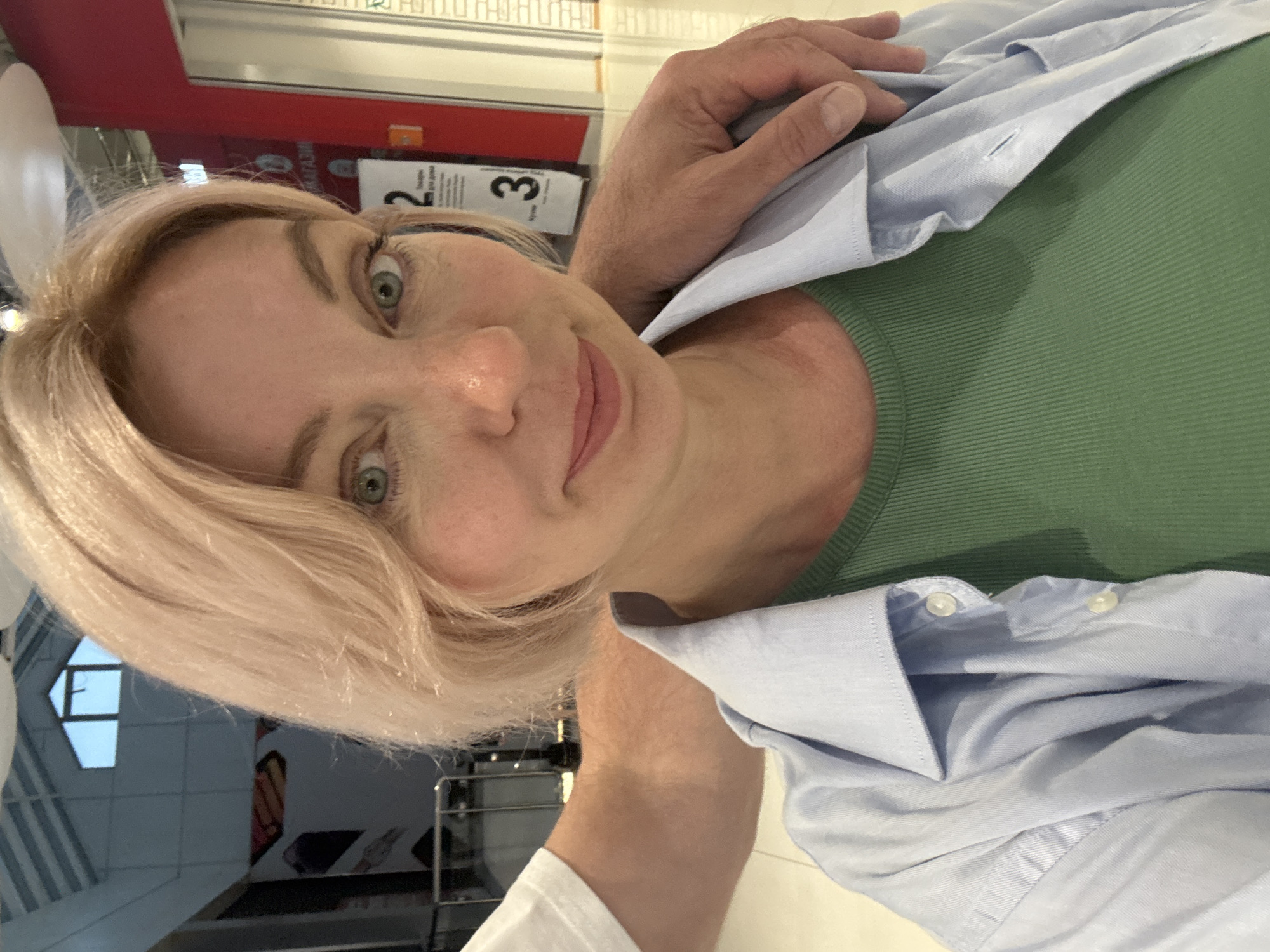 A medium close-up, eye-level selfie captures an adult female and a partial view of an adult male in an indoor retail environment in the city of Sochi, Russia.
The female, positioned centrally, has fair skin, light blonde or strawberry blonde hair styled in a bob, and prominent green eyes. She displays a slight smile and looks directly into the camera. She wears a ribbed dark green crew-neck top beneath an unbuttoned light blue long-sleeved collared shirt.
An adult male's fair-skinned right arm is visible, draped over the female's right shoulder. His hand rests gently on her upper arm/shoulder. He appears to be wearing a white short-sleeved top, with a small portion of the sleeve visible on the left.
The background indicates a brightly lit, modern commercial space, likely a shopping mall or large store. Walls are painted in sections of light grey and red. On the red wall, white Cyrillic text is visible, including "МАГАЗИН" (Store) and "Товары для дома" (Goods for home), accompanied by the large numbers "2" and "3". Overhead, light-colored reflective surfaces and visible structural elements suggest a sloped roof or skylight system. Metal railings are discernible on the left side of the frame. The floor is light-colored and reflective.