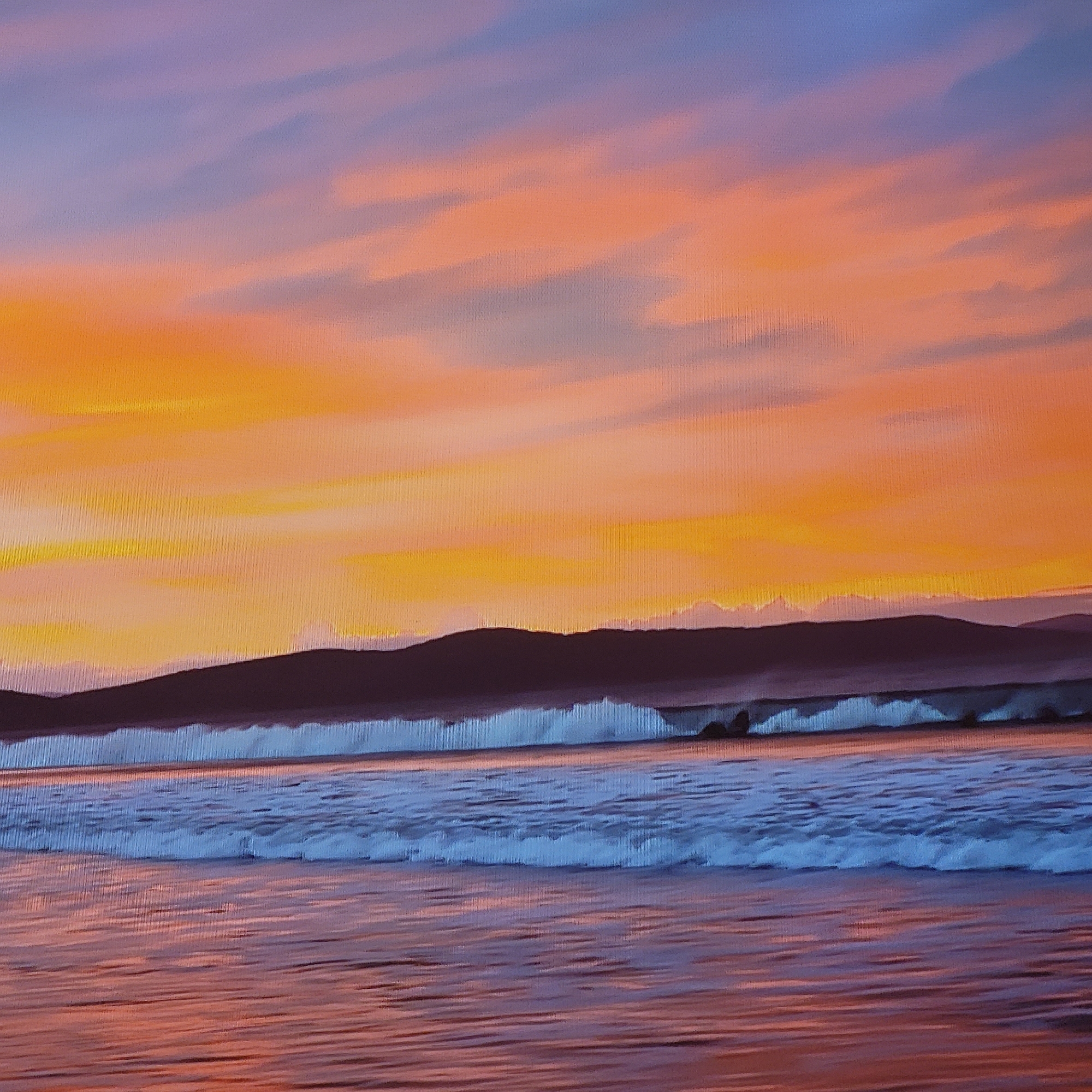 The image depicts a seascape at sunset. The sky is a gradient of colors, transitioning from orange near the horizon to blue higher up, with streaks of pink and gray clouds. Below the sky is a silhouetted mountain range or landmass. In the foreground, waves are breaking on the shore, with white foam crests. The wet sand reflects the colors of the sky. The location is not in Yerevan, Armenia, as it's a landlocked city and does not have a coastline.