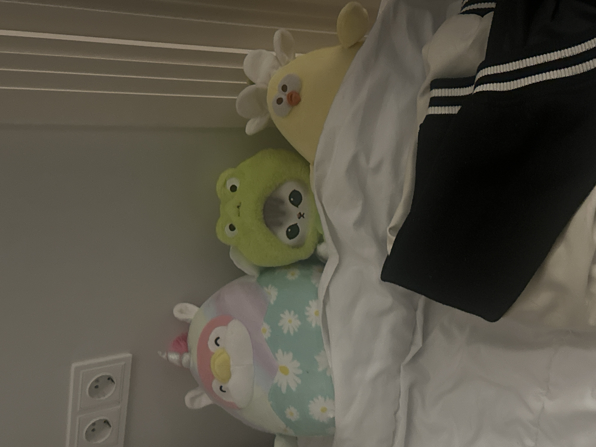The image shows a group of plush toys arranged on a bed. From bottom to top, the toys consist of a pastel-colored unicorn with daisy patterns, a green frog with cartoon eyes, and a yellow chick with a flower on its head. A black garment with white stripes lies on top of the bedding, partially obscuring the toys. The background includes a plain wall with an electrical outlet. The scene is indoors and illuminated with soft lighting. The location is assumed to be in the city of Yerevan, Armenia, based on the presence of a European-style electrical outlet, commonly used there.