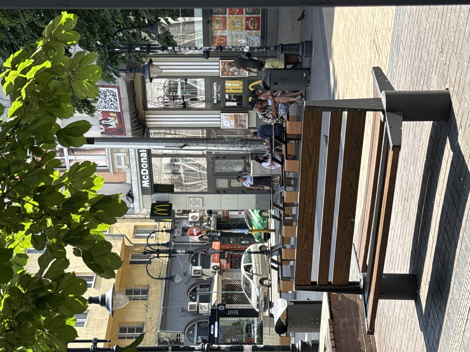 This image shows a street scene with a McDonald's restaurant. There are several people visible, some sitting on a wooden bench in the foreground, and others walking or standing outside the restaurant. The McDonald's sign is clearly visible, as is a "Walk Thru" sign. Other text in the image includes "IRIS STUDIO", "GSM", and what appears to be Bulgarian text such as "СЕРВИЗ" and "ОТАРИУС". The scene suggests a daytime setting with bright sunlight casting shadows. The presence of trees with green leaves indicates it is likely spring or summer. The buildings in the background appear to be shops or businesses. The overall atmosphere seems to be one of everyday activity on a city street. The wooden bench in the foreground is a prominent object, with people using it. A QR code is also visible on a poster attached to a building. There is a green car and a white car parked or driving in the street. The image is taken from a slightly elevated perspective, looking down onto the street and the McDonald's.