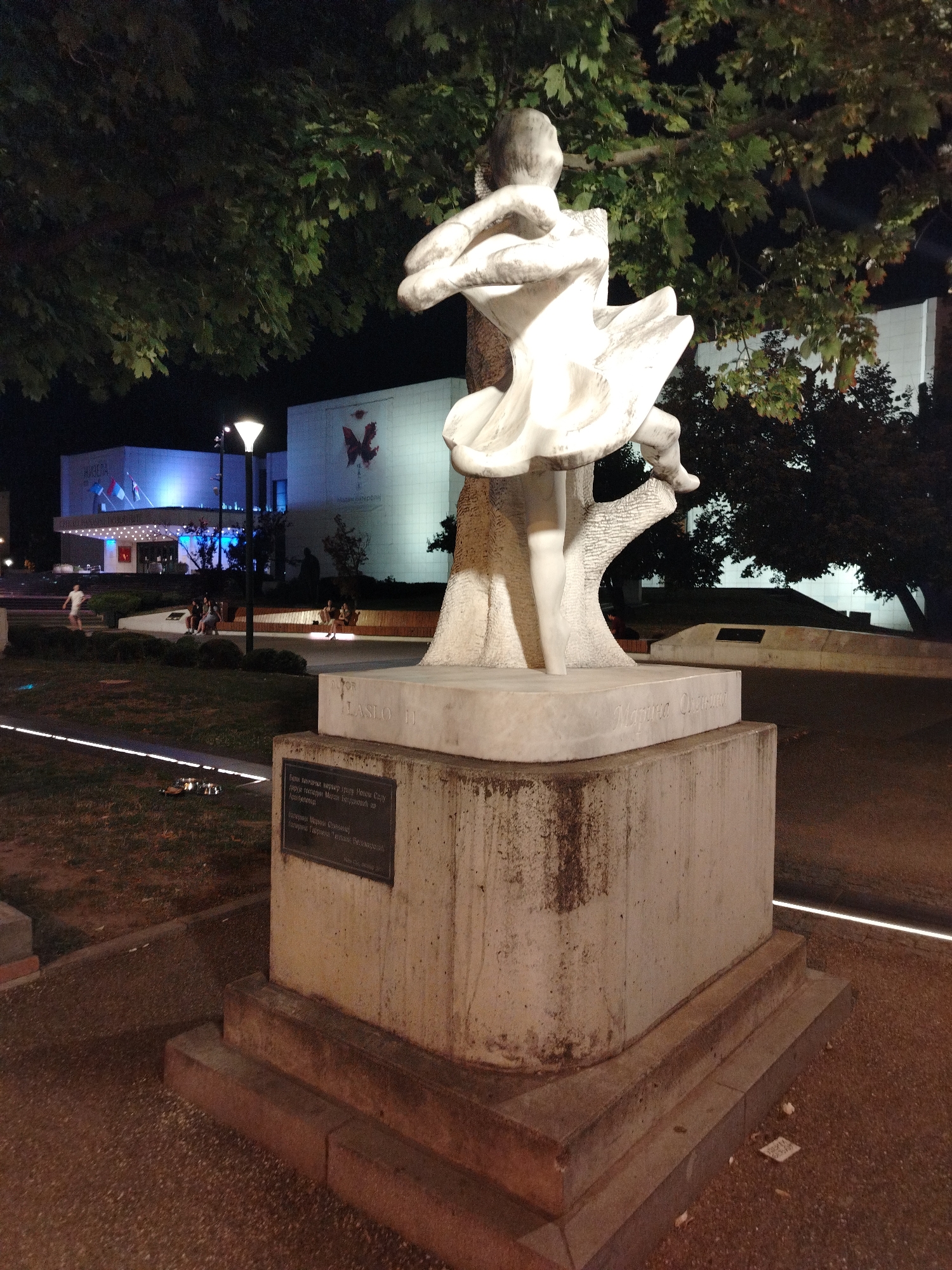 A white marble sculpture of a dancing figure is illuminated at night in front of a building in Novi Sad, Serbia. The sculpture, a woman in a flowing dress, is on a concrete pedestal. A bronze plaque is attached to the pedestal. The building behind the sculpture is lit with blue lights and features a red butterfly graphic. Several people are seated on benches in the background.