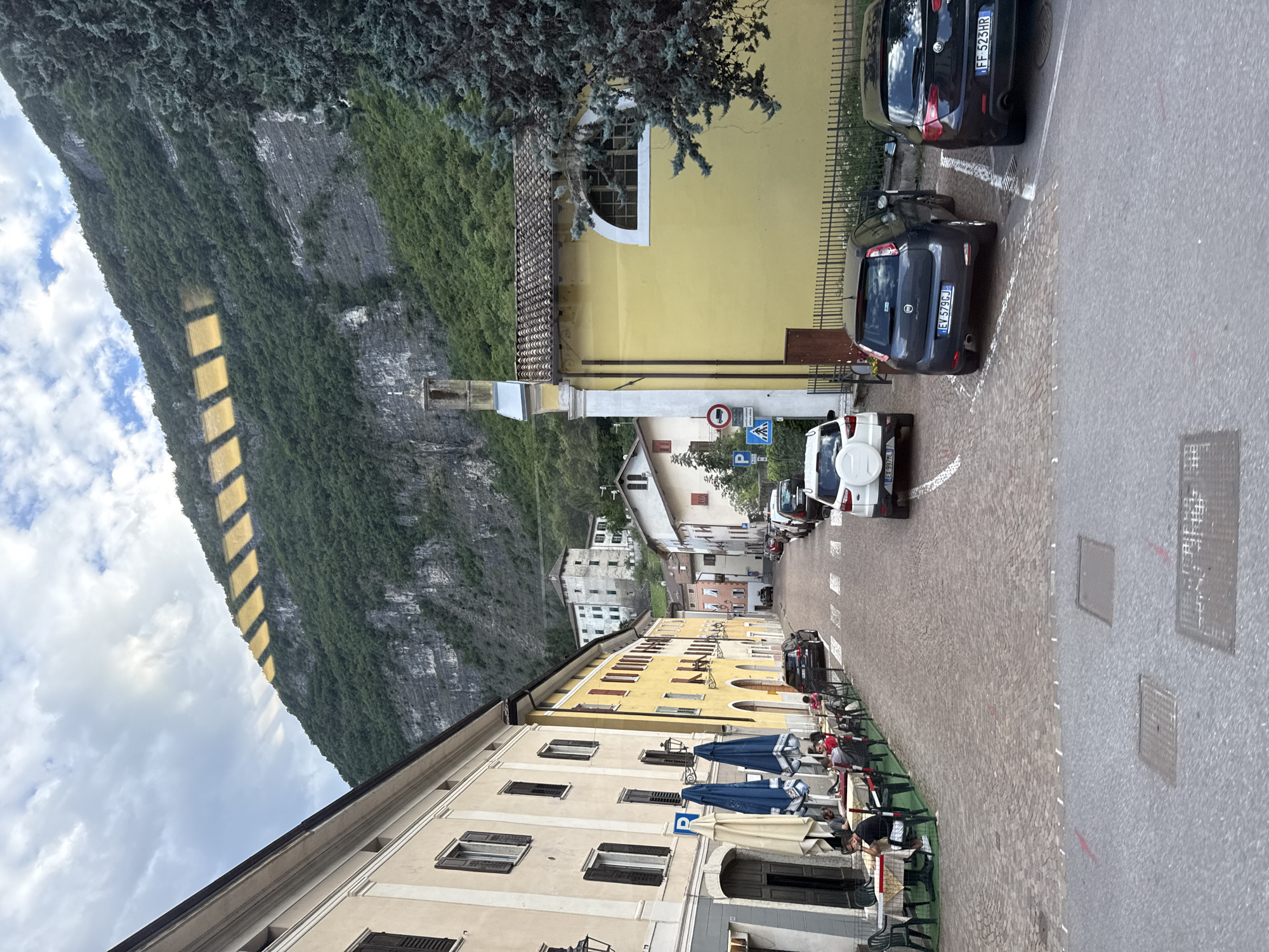 The image depicts a street scene in Mezzolombardo, Italy, likely during daylight hours. The street is paved with cobblestones and lined with buildings on both sides. The architecture suggests a European town, with multi-story buildings featuring traditional windows and shutters. On the left, a building with a light-colored facade has tables and chairs set up outside, indicating a cafe or restaurant, with several patrons seated. Blue and white umbrellas provide shade over the outdoor seating.
The street slopes gently upwards into the distance. Several cars are parked along the right side of the street and some are in motion. A white SUV is parked on the right, followed by a dark gray hatchback and a black sedan. Further up the street, more parked cars are visible.
In the background, a large, tree-covered mountain rises steeply. The sunlight creates a pattern of rectangular shapes, possibly due to reflections or sunlight filtering through something, on the upper part of the mountain. A yellow building with a tiled roof and a tall, slender bell tower is prominent on the right side of the street. A few traffic signs are visible, including a pedestrian crossing sign and a no-trucks sign. The sky is partly cloudy, with patches of blue visible.