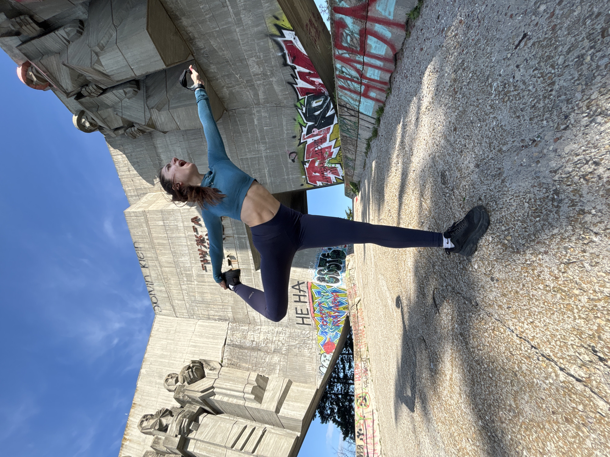 A woman is captured mid-air, performing a split jump in front of the Monument of the Bulgarian-Soviet Friendship in Varna, Bulgaria. She wears a blue long-sleeved crop top and dark leggings with black running shoes. Her arms are extended, one upward towards the monument and the other backward. Her head is tilted back, and her mouth is open in what appears to be a shout. The monument features concrete reliefs of figures. Graffiti is visible on the concrete walls. A concrete ground surface is visible, casting shadows. Blue sky is visible above.