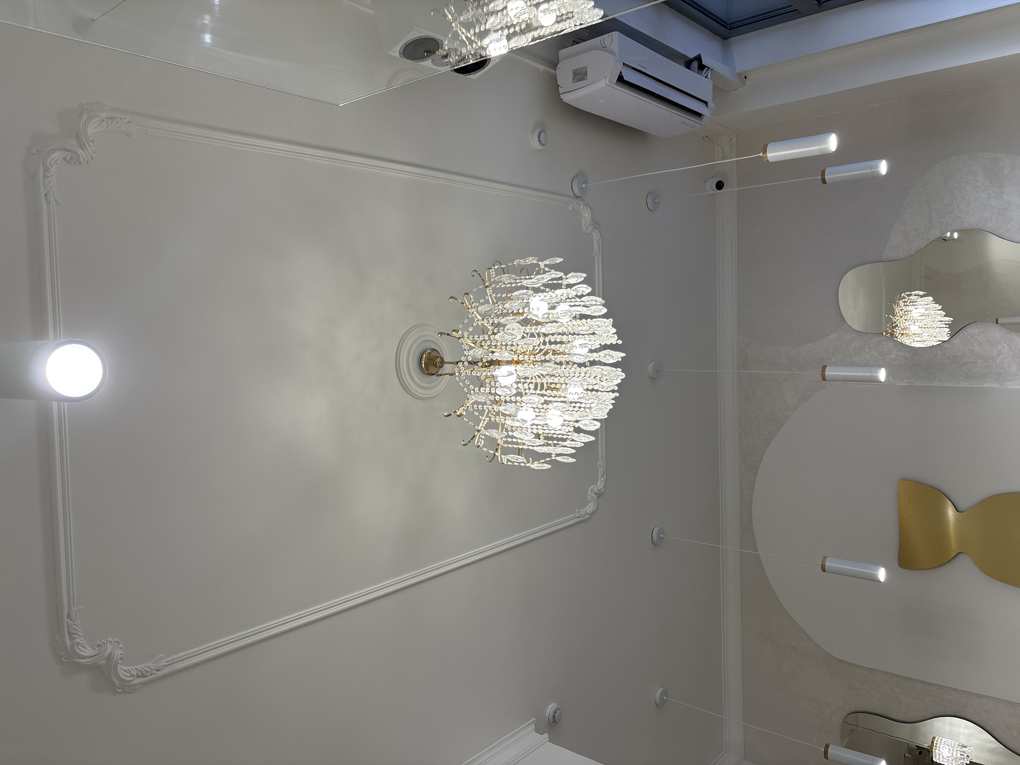 This upward-facing image showcases a decorative interior ceiling and upper wall in Krasnoyarsk, Russia, blending classical and modern design elements.
At the center, a large, ornate crystal chandelier with numerous shimmering facets hangs from the ceiling, serving as a prominent light source. Surrounding it is a rectangular frame of elaborate white classical molding, adding a traditional architectural detail to the ceiling. A bright, circular spotlight is visible in the upper left corner, illuminating the space.
The right side of the room features contemporary elements. Several white, cylindrical hanging lights extend horizontally from the wall on thin wires, casting focused light. Below these, a series of artfully shaped mirrors with irregular, wavy edges are mounted on the wall; one mirror distinctly reflects a portion of the chandelier. A distinctive golden, abstract, bow-tie or fish-shaped decoration is positioned on the wall.
High on the upper right, a white air conditioning unit bearing the "Haier" brand name is visible. Nearby, a small black security camera is mounted on the wall. Various white circular components, likely smoke detectors or sensors, are distributed across the ceiling and walls. Reflections of overhead structures or lights are apparent in a glossy surface at the very top of the image. The entire space is well-lit by artificial sources, creating a bright and sophisticated ambiance.