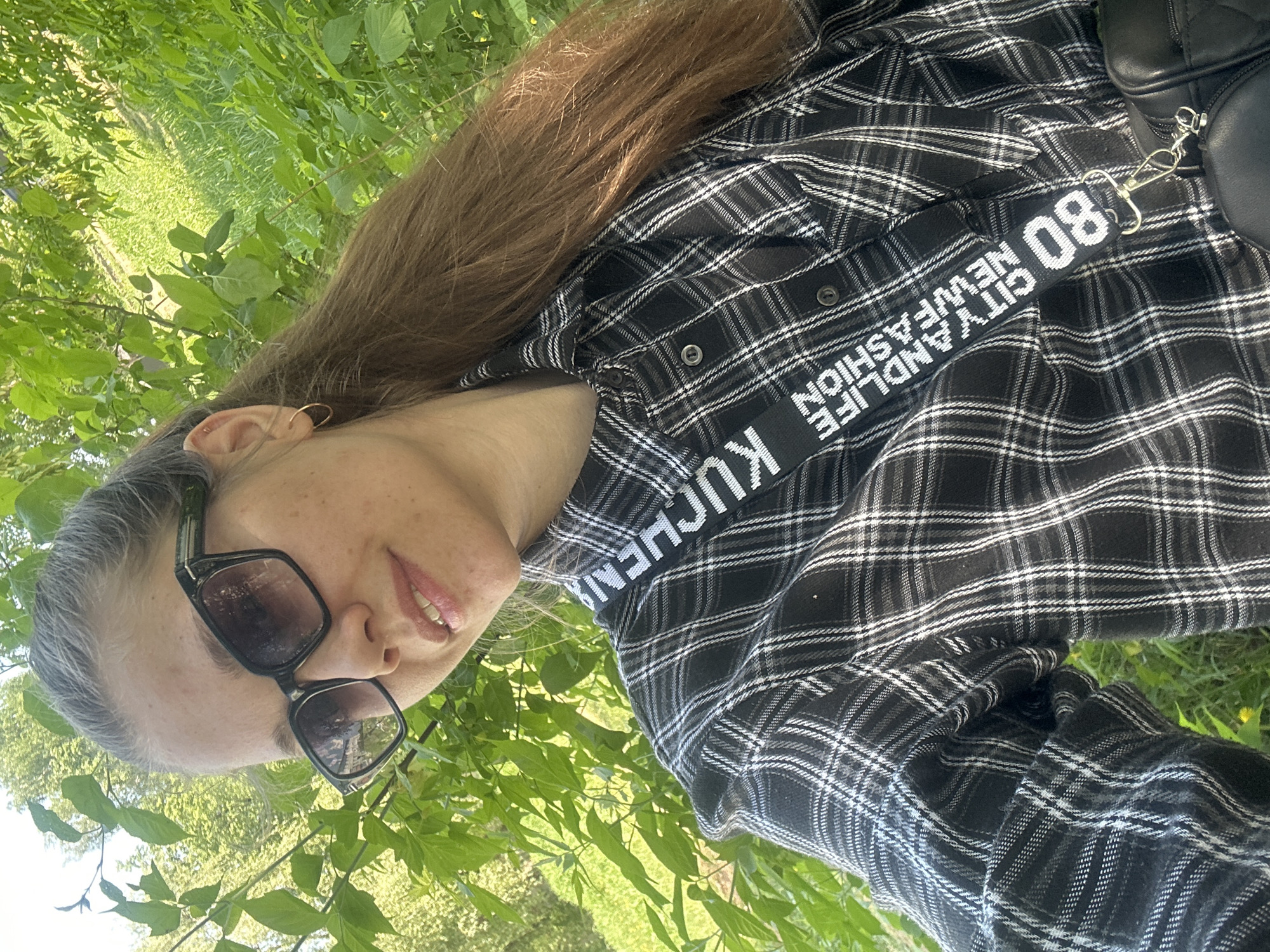 A woman wearing sunglasses and a black-and-white plaid shirt poses for a selfie outdoors. She is positioned against a backdrop of green foliage, suggesting a park or other natural setting. The text "80 CITYANDLIFE NEWFASHION" and "KUCHEN" are visible on her shirt. It is not possible to determine the location of the city of Chaikovsky, Russia, from the image alone.