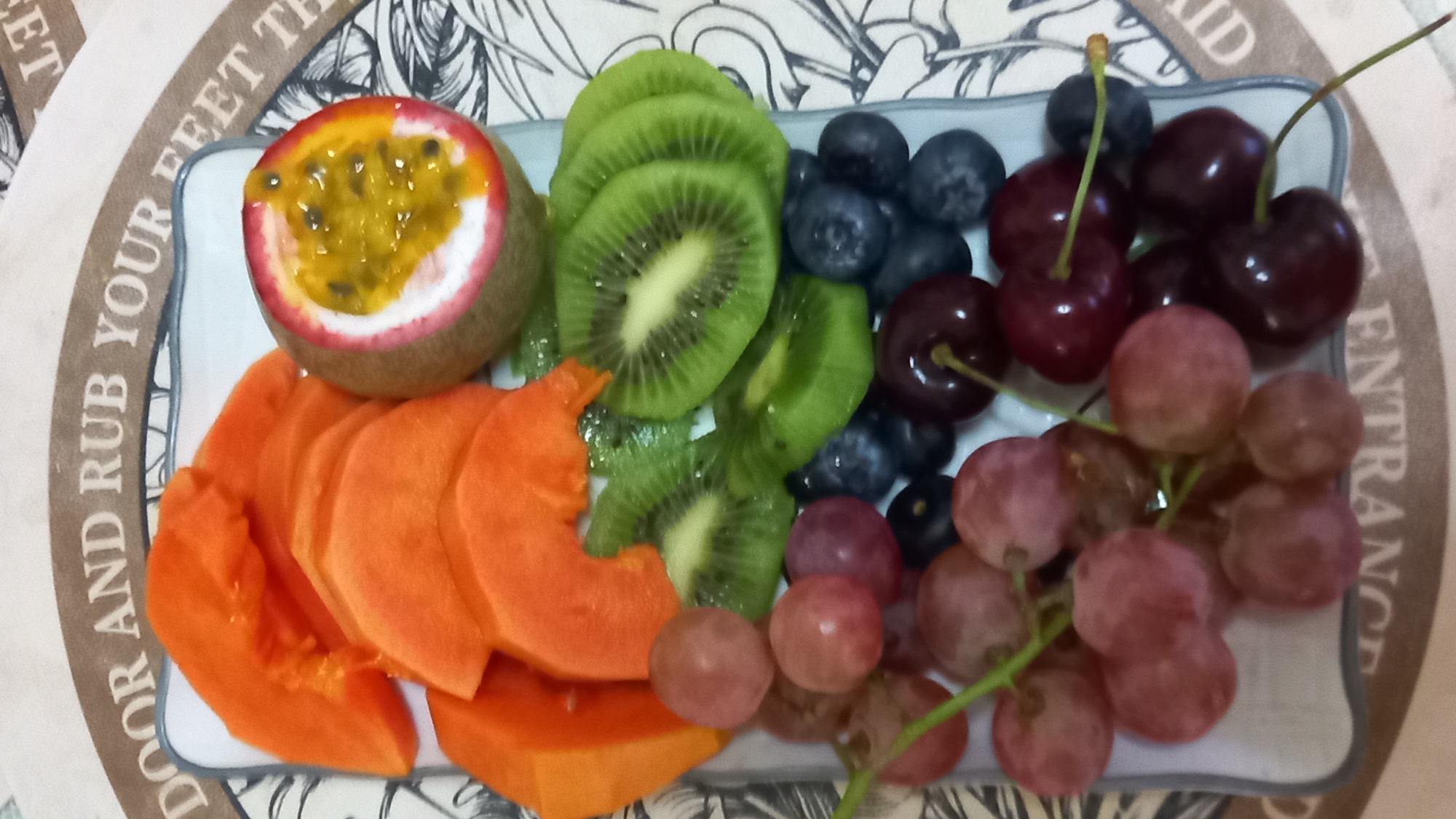 The image shows a close-up, overhead shot of a platter of assorted fruits, likely in an indoor setting, possibly a restaurant or a home in Koh Phangan District, Thailand. The platter, which is white and rectangular with a blue trim, contains sliced papaya, a halved passion fruit, sliced kiwi, red grapes, blueberries, and cherries. The fruits are arranged artfully on the platter. In the background, a circular mat with text is visible, with the words "DOOR AND RUB YOUR FEET TH" partially legible. The overall impression is one of a fresh and vibrant fruit selection. There are no people or animals visible in the image.