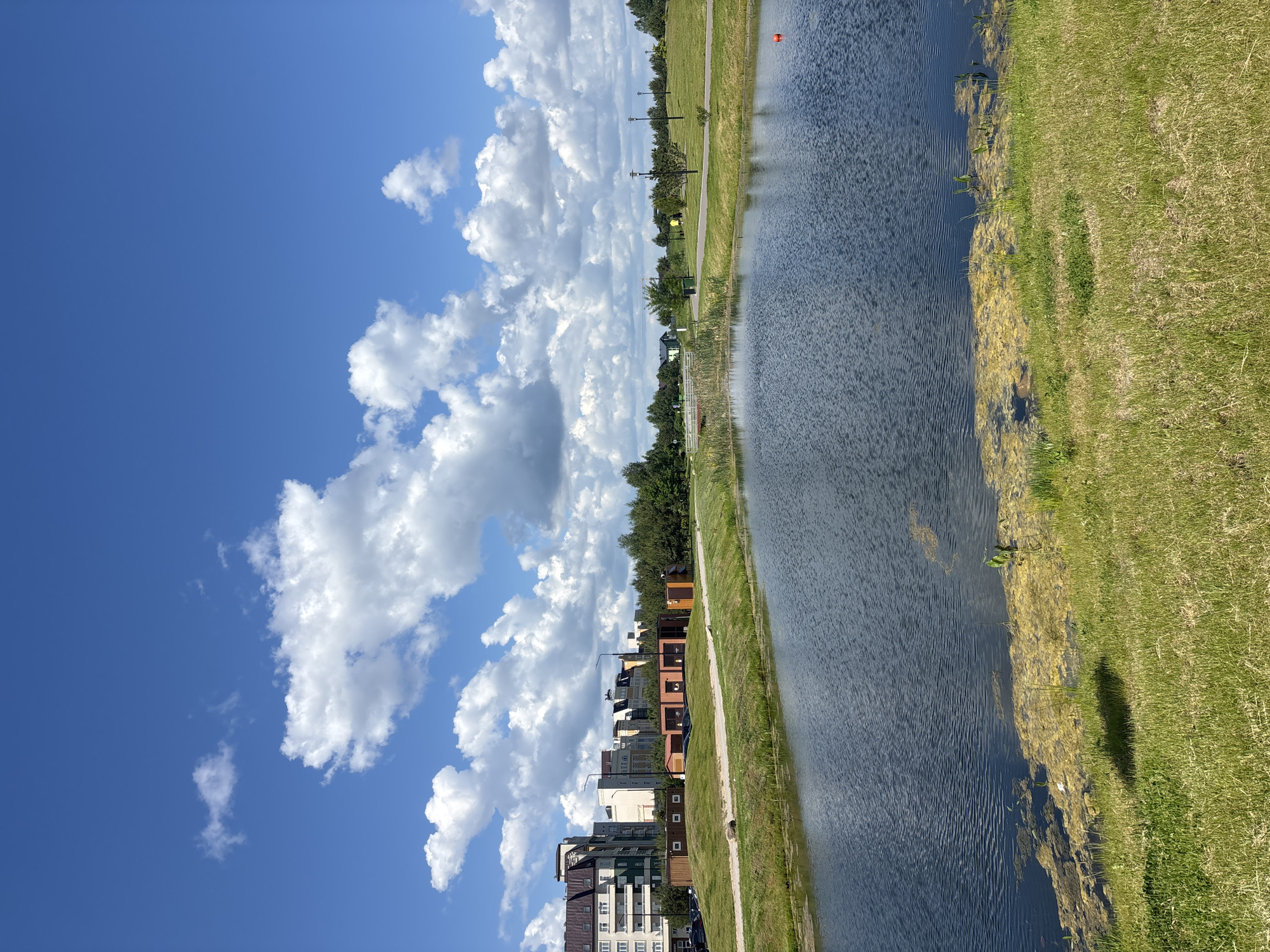 A vertical-oriented outdoor scene in Varaksino, Russia, features a body of water (likely a canal or lake) to the right, with a grassy bank and some aquatic vegetation along its edge. To the left of the water is a path or walkway, beyond which are buildings of varying heights and architectural styles. A clear blue sky with scattered, puffy white clouds occupies the upper left portion of the frame. The light suggests a sunny day.