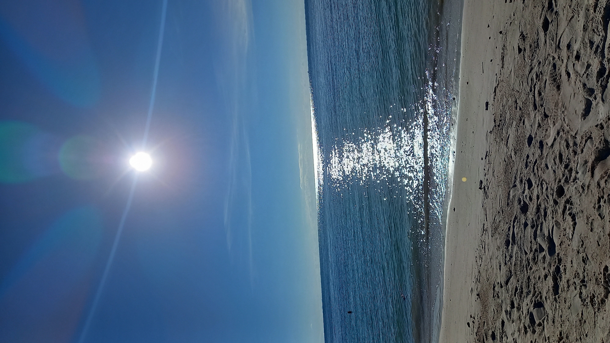 This is a vertical photograph of a sun-drenched beach. The sun is high in the clear blue sky, casting a bright glare on the water. The water is a deep blue and reflects the sunlight, creating a shimmering effect. The beach is sandy and appears to be uncrowded. The sand is light brown and textured with footprints. The edge of the water gently laps at the sand. The overall impression is one of a peaceful and beautiful beach scene on a sunny day. The photograph was taken in Ko Pha Ngan, Thailand.