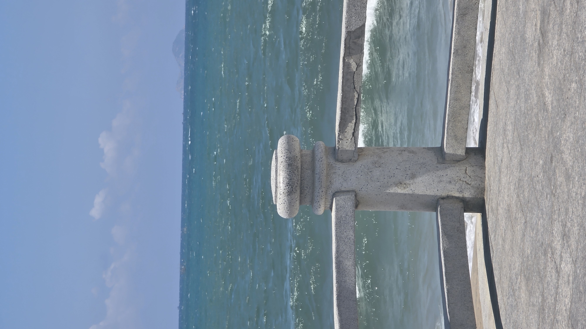 The image shows a close-up view of a concrete pier railing with the ocean and sky in the background. The railing is light gray and appears weathered, with a decorative concrete cap on the post. The ocean is turquoise, with visible waves and white foam near the pier. The sky is blue with scattered white clouds. The land in the background indicates the city of Nha Trang Ward, Vietnam.
