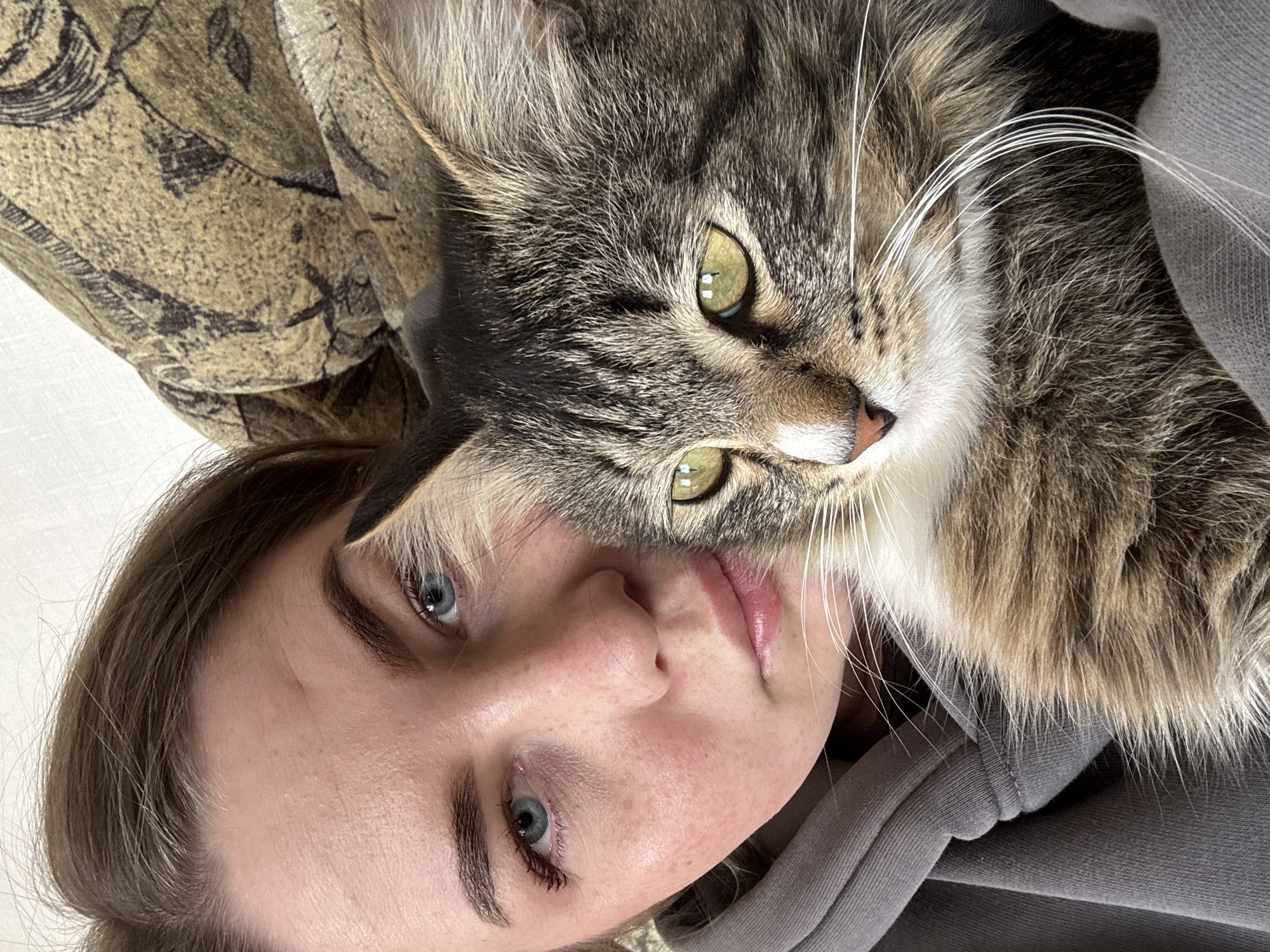 A close-up photograph shows a woman with fair skin and blue eyes laying on a patterned surface. She is positioned to the left, and a tabby cat with green eyes is resting on her face. The woman is wearing a gray hooded garment. The background includes a patterned fabric with a gold and black design. There is no evidence to suggest the location is in Гаревая, Russia.