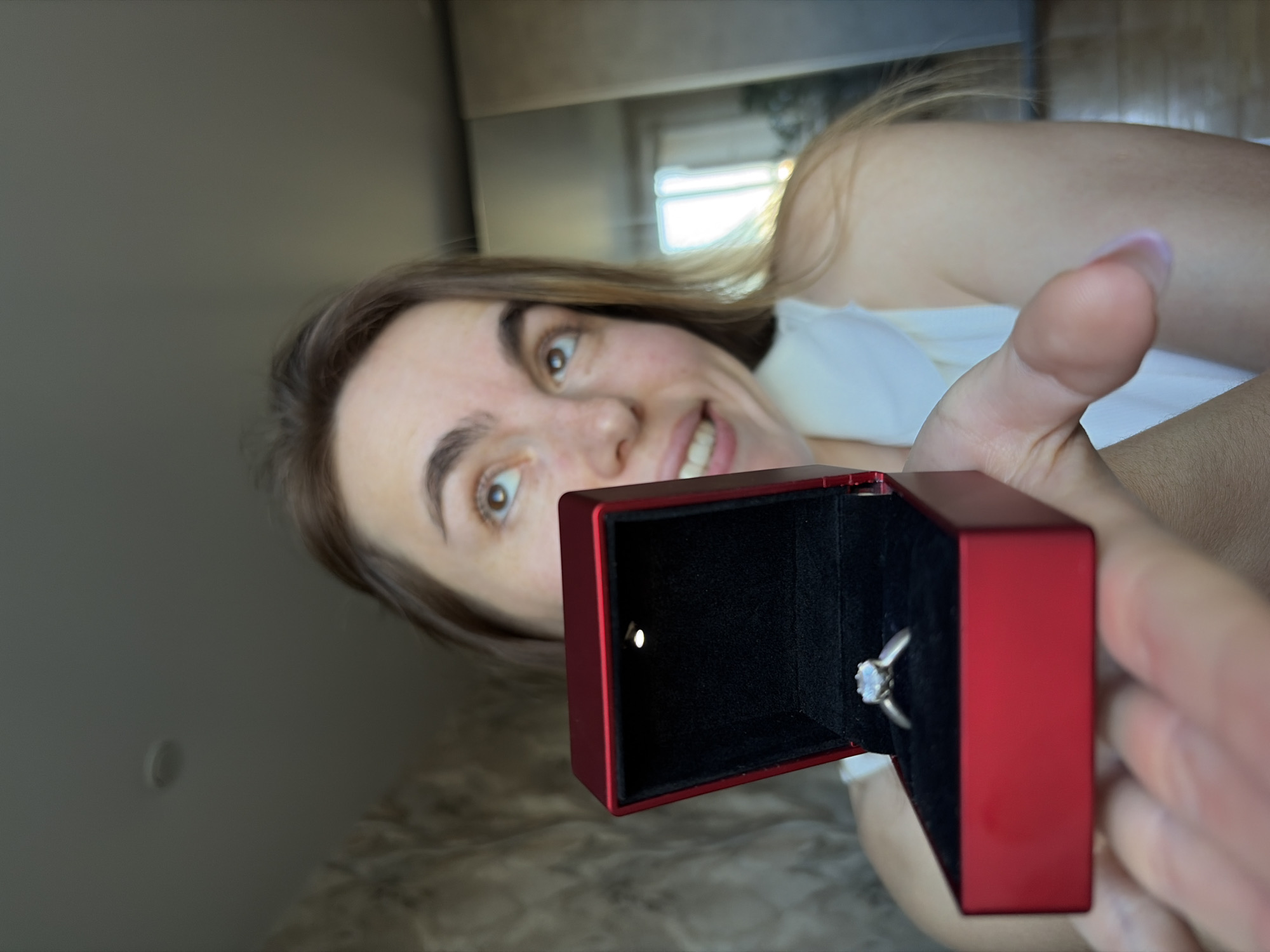 A woman holds open a red ring box with a ring inside. The woman is fair-skinned with light brown hair and brown eyes. She is smiling and looking at the camera. The box is lit from within, highlighting the ring. The background is blurry but appears to be an interior space. There is no indication of the image location within Izhevsk, Russia.