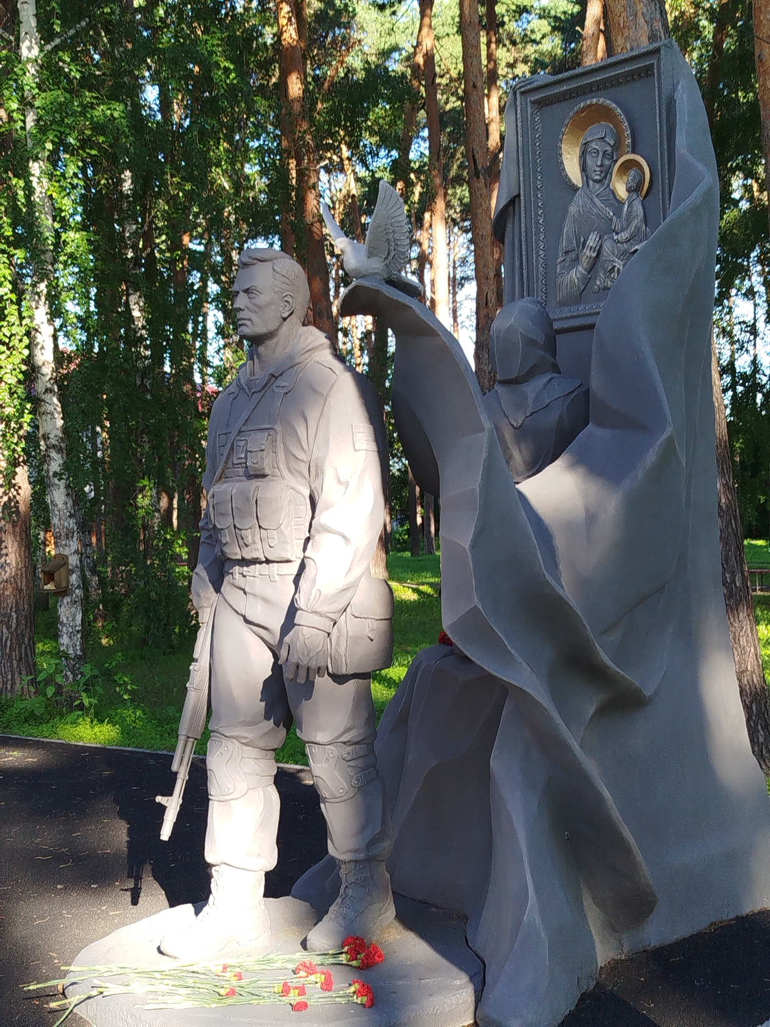 This is a photograph of a memorial sculpture located in Zavodoukovsk, Russia. The sculpture depicts a soldier in military attire, standing with a rifle at his side. To the right of the soldier is a large, abstract stone structure with a religious icon of the Virgin Mary and child embedded in it. A dove is perched on the abstract structure above the soldier. Red carnations have been placed at the base of the sculpture. The background consists of trees and greenery, indicating an outdoor setting, likely a park or memorial site. The lighting suggests it is daytime, with sunlight casting shadows on the sculpture.
