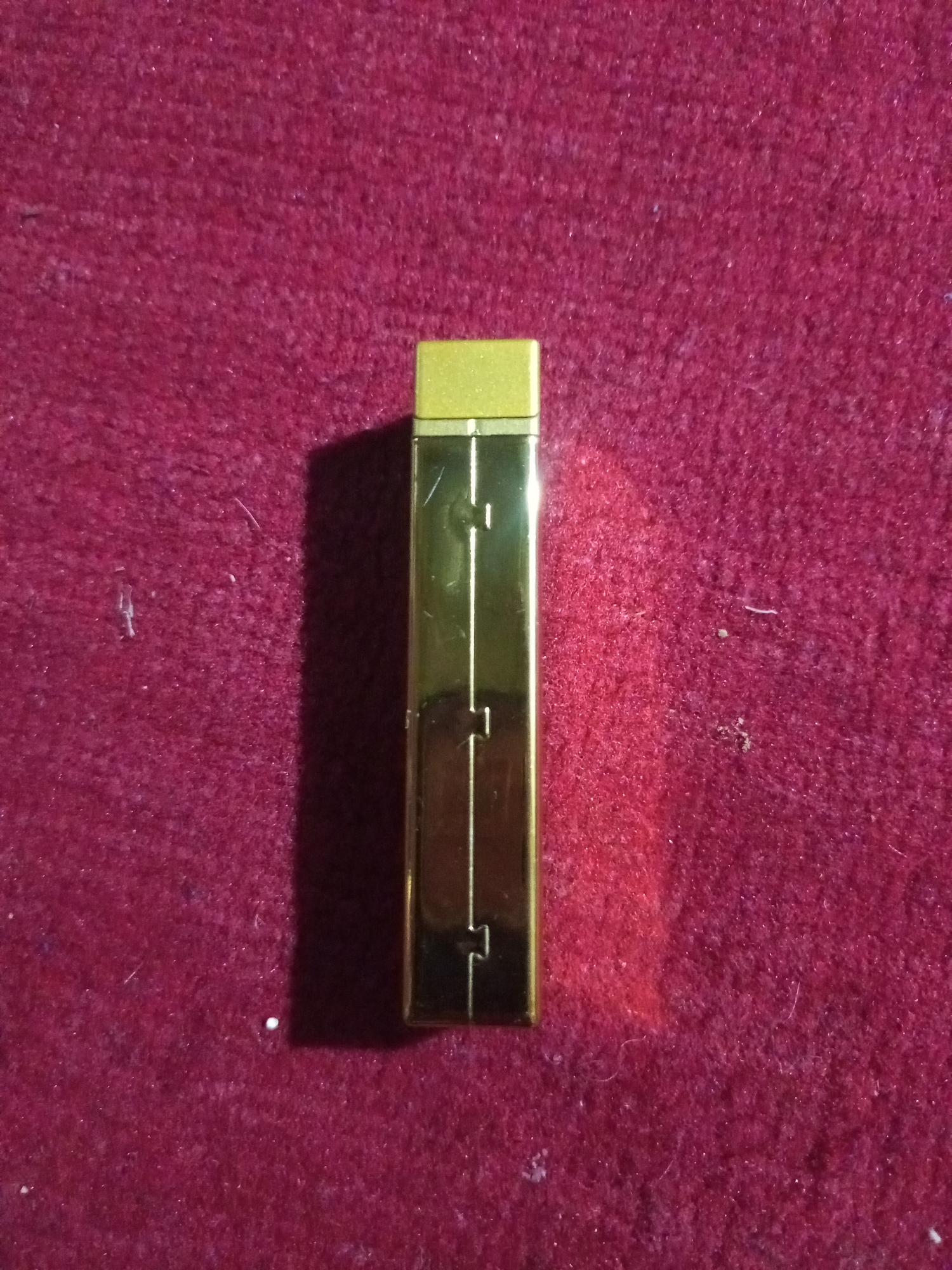 A gold-colored rectangular object, possibly a lighter or a small electronics device, is positioned vertically in the center of a dark red, textured surface. The object has a metallic sheen and features a thin, recessed line running down its center, with horizontal indentations at the top and bottom of this line. The surface beneath is a deep red, appearing to be made of a soft, possibly fabric material with a slightly fuzzy texture. The lighting casts a shadow to the right of the object, indicating a light source from the left. The image does not contain any information to place it within the city of Lhokseumawe, Indonesia.
