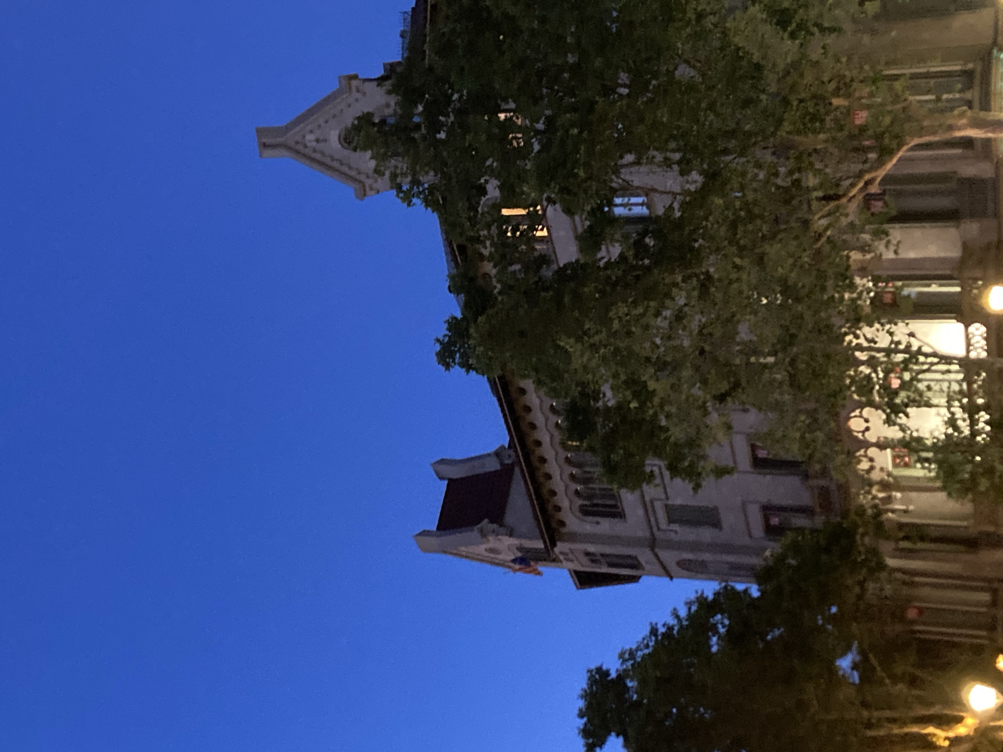 The image depicts an upward-angle view of a building facade in Barcelona, Spain, at twilight. The sky is a deep, clear blue, suggesting fair weather. Green foliage from trees partially obscures the building, which appears to be made of light-colored stone with ornate architectural details. Windows are visible, some illuminated from within, indicating that the building is occupied. The building's roofline is visible, with a red-tiled section. Streetlights are also visible at the bottom right of the image, casting a warm glow, further confirming it is evening. The perspective is from street level, looking up towards the upper stories of the building. There is no visible text in the image. The scene evokes a quiet urban evening atmosphere.