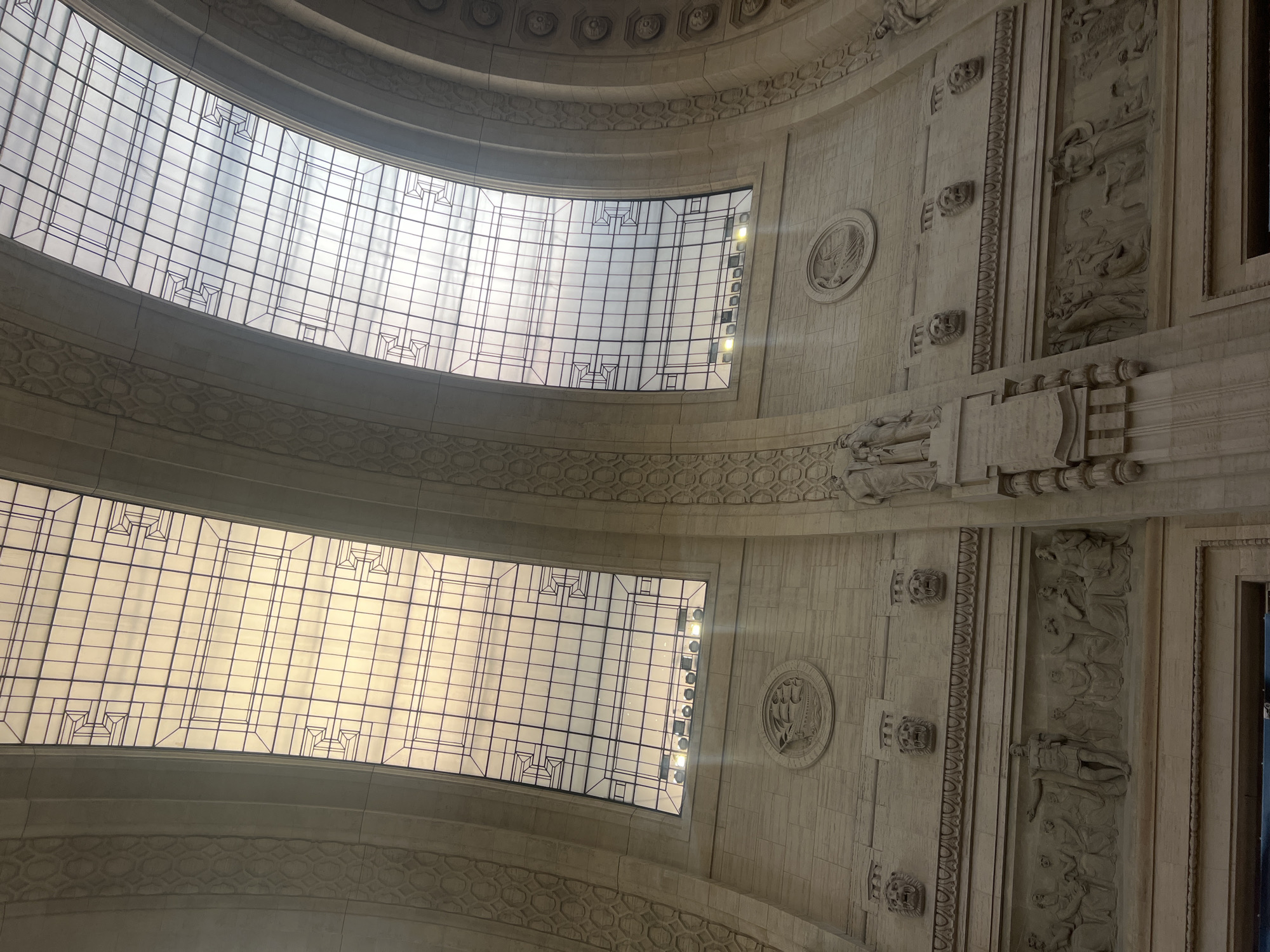The image shows the interior of the Galleria Vittorio Emanuele II in Milan, Italy, looking upwards. The scene features the glass-domed ceiling with its intricate grid pattern. The walls are made of stone and ornamented with bas-relief sculptures, medallions, and repeated lion head motifs. Sunlight is filtering through the glass panels.