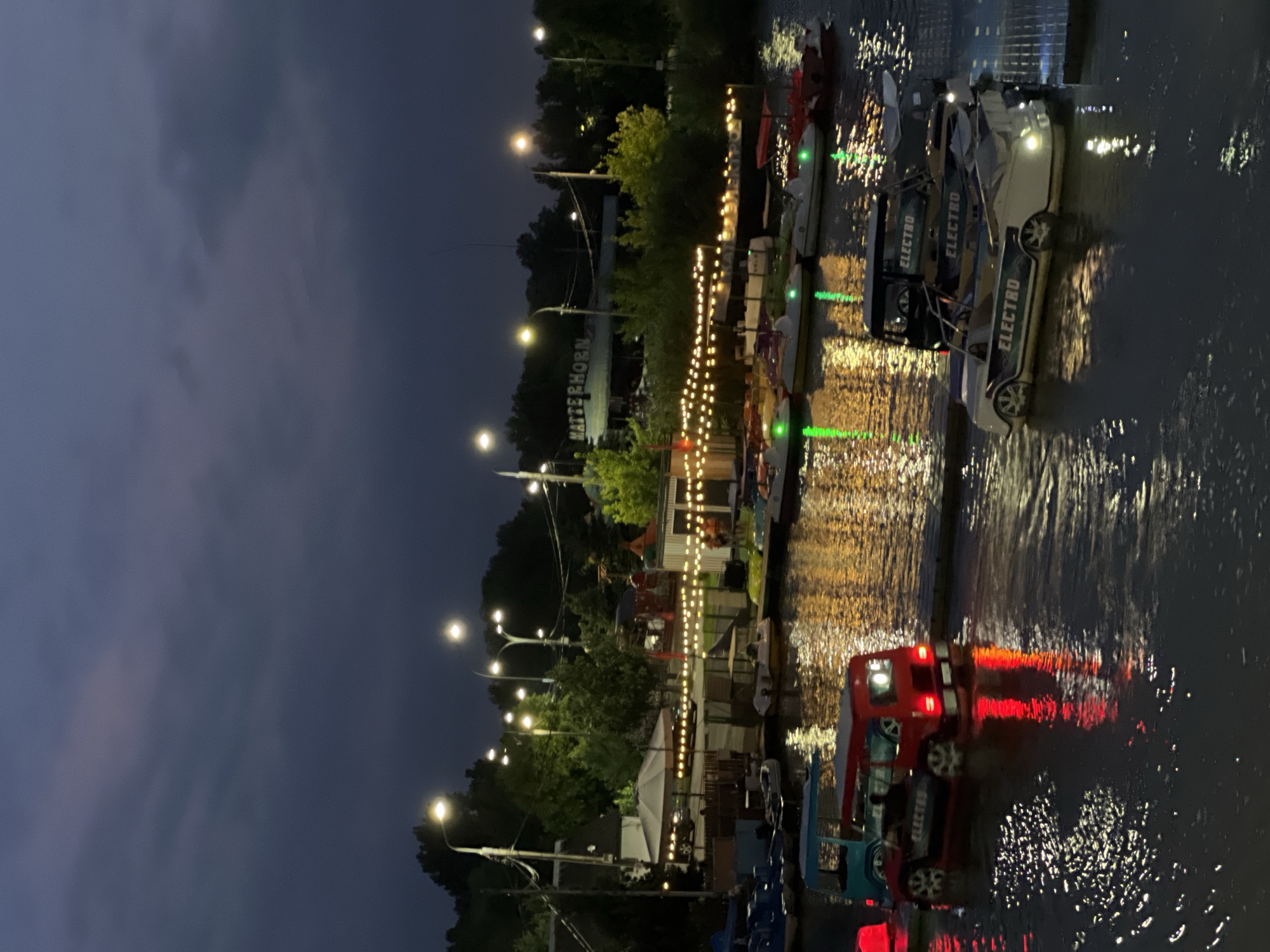 An outdoor scene at dusk shows a body of water with several small pedal boats and electric boats docked or on the water. The boats are various colors, including red, blue, white, and yellow, and some have lights. In the background, there are trees, streetlights, and buildings, including one with a sign that says "MATTERHORN." There are strings of decorative lights draped across some areas, and a large umbrella is visible. Several people are present near the buildings and boats. The water reflects the lights from the boats and surrounding area, creating a shimmering effect. The setting appears to be a park or recreational area.