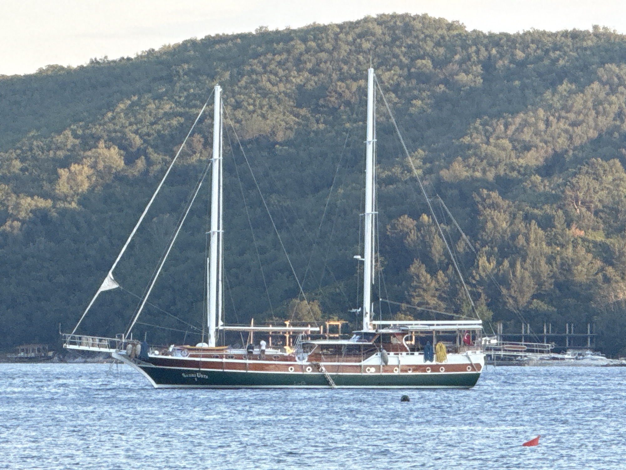 A medium shot depicts a two-masted yacht floating on calm water. The yacht is primarily green and brown, with a white deck and white masts and rigging. A small, triangular sail is partially unfurled on the forward mast. The yacht's name, "Siren Usta," is visible on its hull.
In the background, a heavily forested hillside rises, filling the upper portion of the frame. A small structure, possibly a dock or pier, is visible in the distance behind the yacht. A small red marker buoy floats in the water to the lower right of the yacht.
The location is identified as the city of Budva, Montenegro. The overall scene is one of serenity and maritime activity.