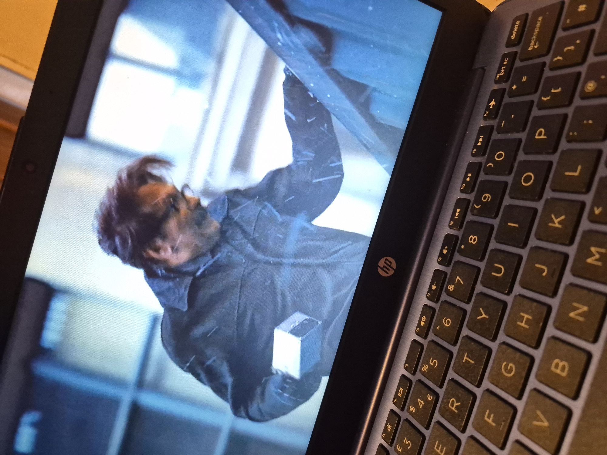 The image shows a laptop screen displaying a scene from a film or television show. A man, appearing to be in his 40s or 50s with dark hair and glasses, is wearing a dark jacket and holding a small, light-colored rectangular object in his left hand. He is positioned in the foreground of the screen, with a blurry, blue-toned background suggesting an outdoor urban environment, possibly in a city. Snow or rain appears to be falling. The laptop itself is partially visible in the lower portion of the image, with a keyboard and the HP logo clearly identifiable. The perspective suggests the image was taken from a slightly elevated angle, looking down at the laptop. There is no information in the image to indicate a specific location in the city of Sandwell, United Kingdom.
