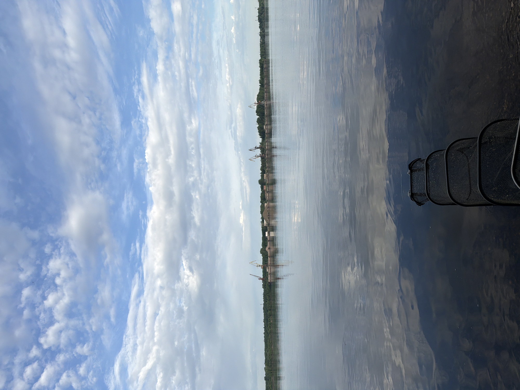 The image depicts a body of water, possibly the Kama River, in the city of Chaikovsky, Russia. The sky is partly cloudy with blue patches. The shoreline features vegetation and industrial structures, including cranes, indicative of port or dock facilities. A fishing net is partially visible in the foreground on the right. The water reflects the sky and the shoreline.