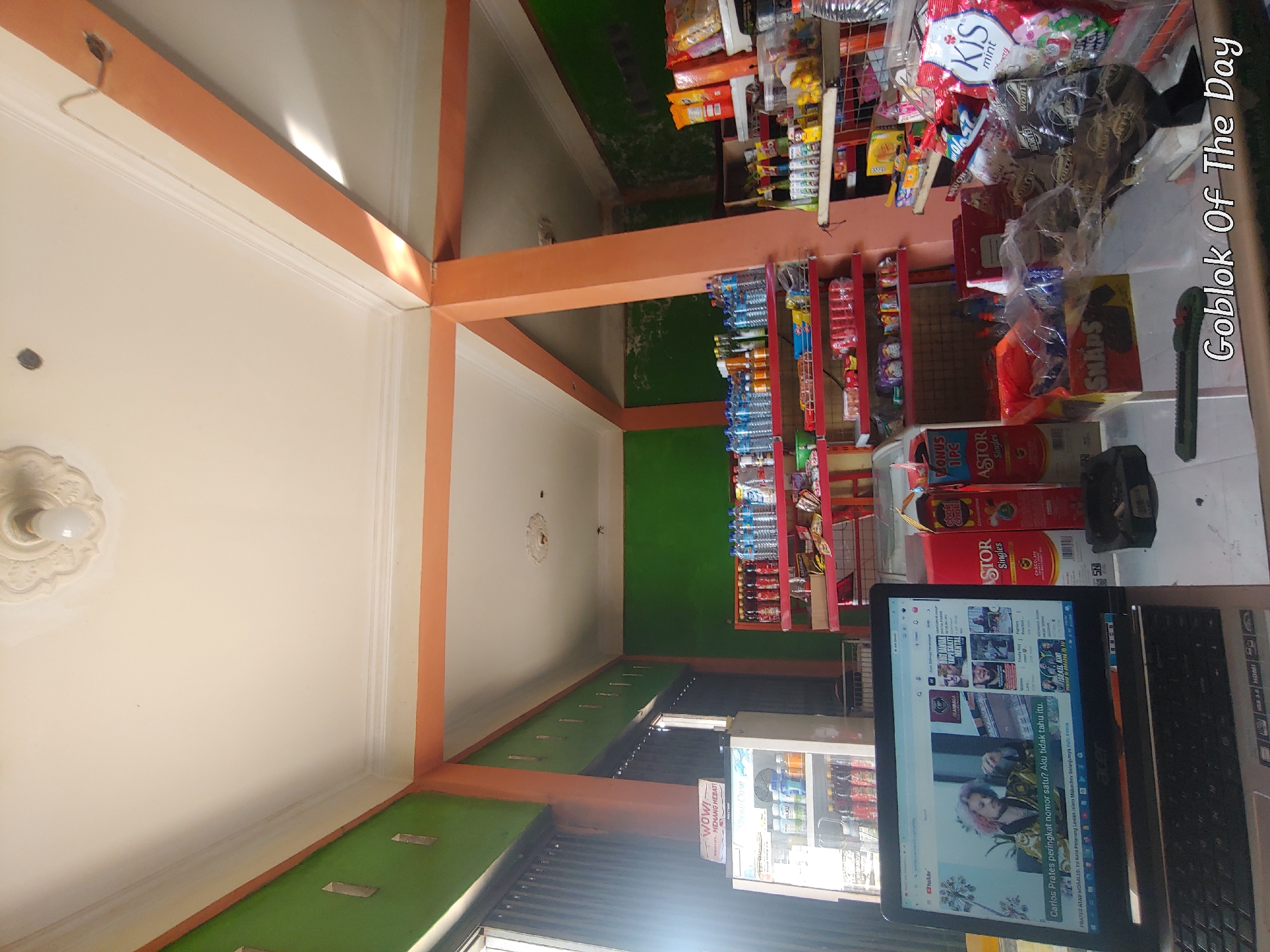 The image depicts the interior of a small convenience store, likely a warung, in Kembang Kerang, Indonesia. The scene is captured from a low angle, looking upwards and across the store. In the foreground, a laptop is open, displaying a YouTube video with text in Indonesian. To the right of the laptop are several snack items, including boxes of "Astor Singles" and "Choki Choki". Further back, shelves are stocked with various beverages, snacks, and packaged goods. Prominent are bottles of water and soft drinks. The walls of the store are painted green and orange, with an orange beam supporting the ceiling. A small refrigerator is visible on the left, containing more drinks. The lighting suggests it is daytime, with some natural light entering from the left. The overall impression is of a modest, well-stocked local shop.