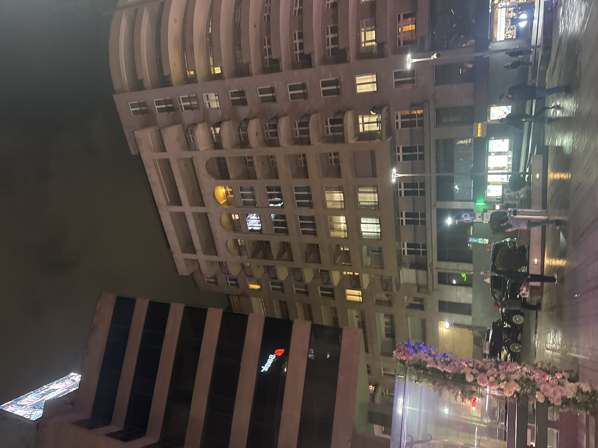 The image depicts a night scene in Yerevan, Armenia. The foreground features a modern building with dark vertical stripes and a sign with a red heart logo. A building adorned with ornate balconies and multiple illuminated windows occupies the upper section of the image. A partially visible pedestrian walkway shows people walking. Cars and decorative floral arrangements are visible on the right side of the image. The light levels indicate that the image was taken at night.