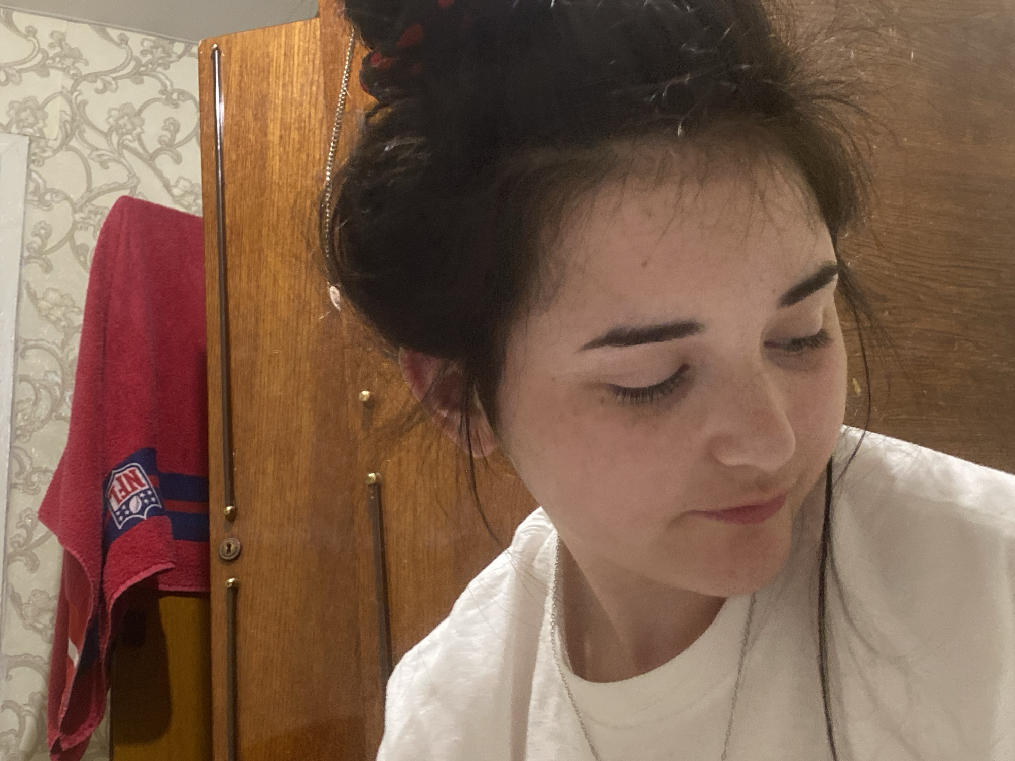 A close-up shot features a woman with dark hair pulled up. She is wearing a white t-shirt and a silver necklace. Her eyes are downcast and she has an expression of introspection. In the background, there is a wooden wardrobe with vertical metal handles and a red towel with a logo. The wallpaper behind the wardrobe is patterned with floral designs. The location is likely indoors. There is no confirmed evidence that this image was taken in the city of Poltava, Ukraine.